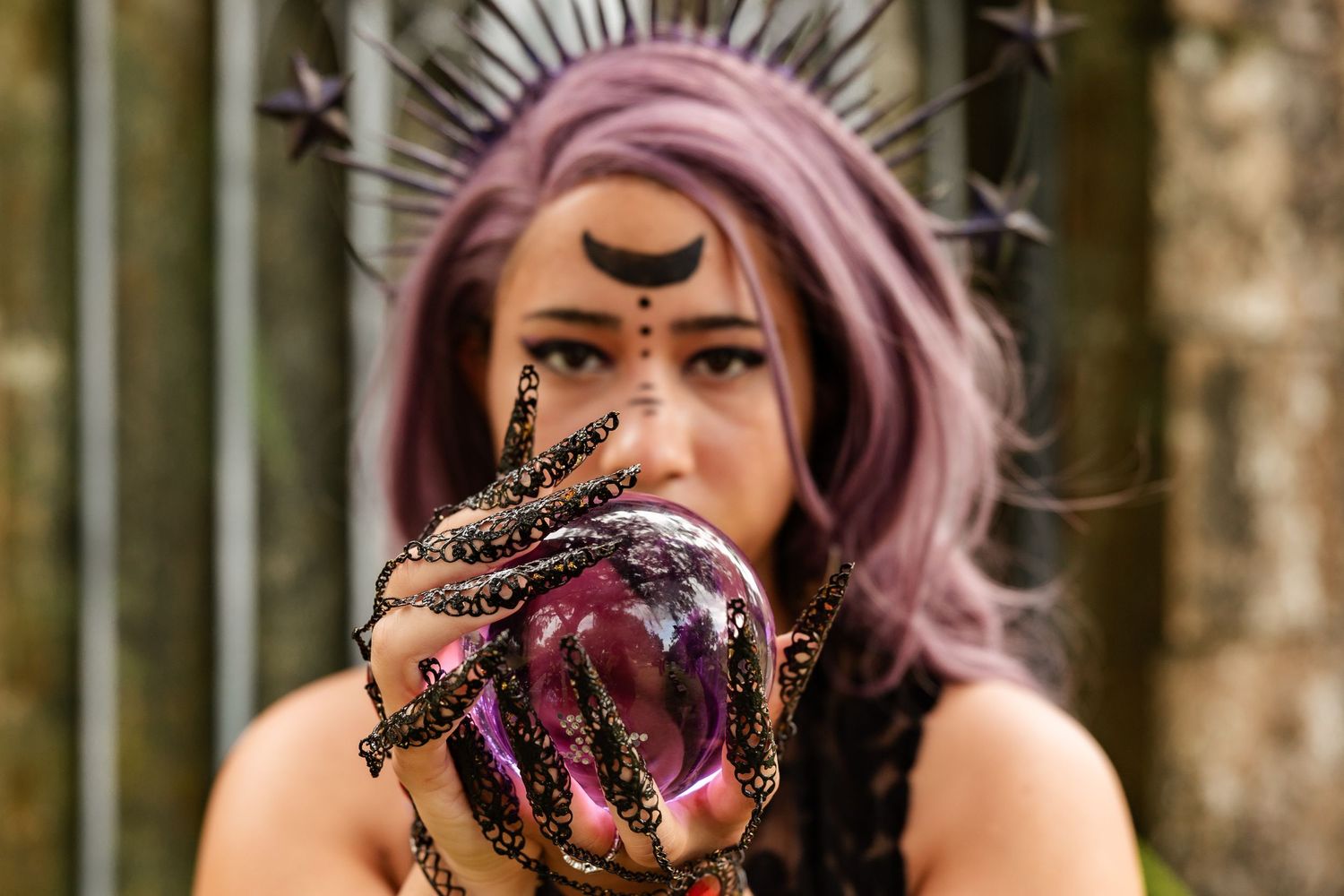 Woman with purple hair and moon face paint holds a glowing crystal ball with ornate black claw rings.