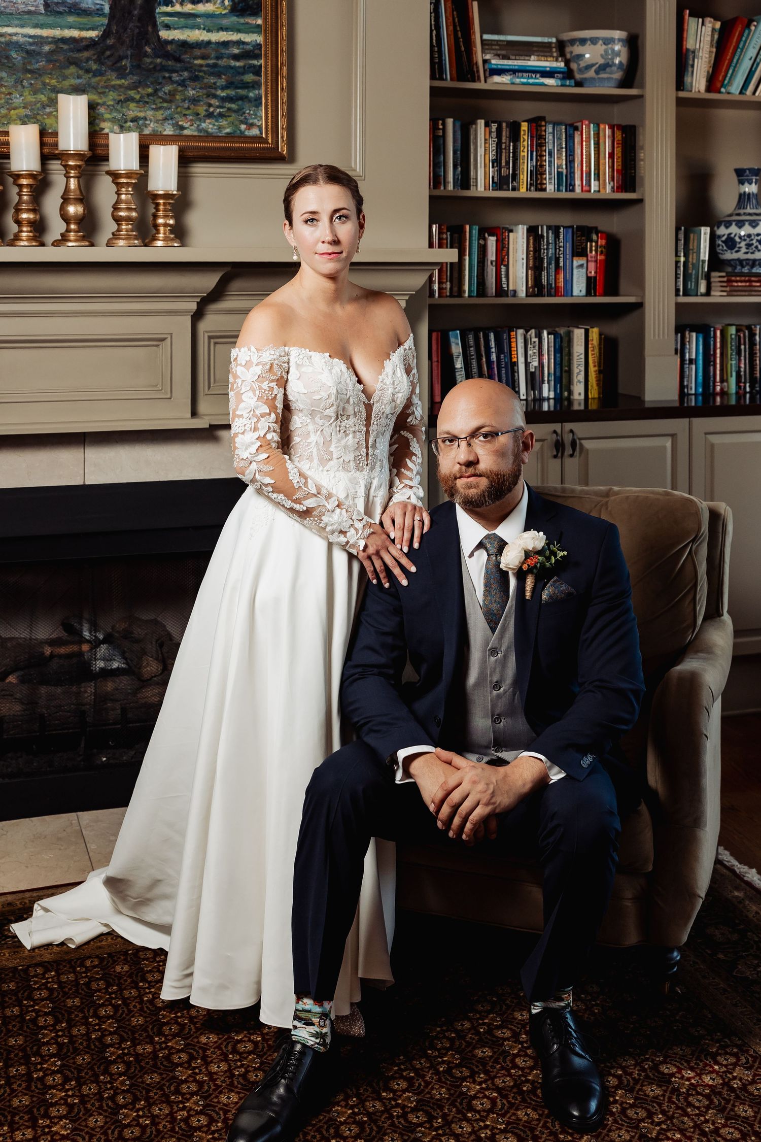 Bride stands beside seated groom in navy suit posing for formal portrait by fireplace and bookshelf.