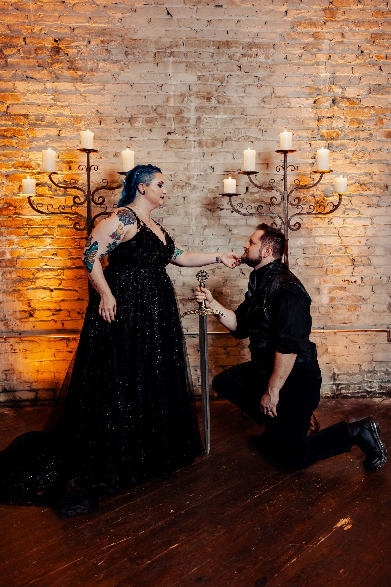 Gothic groom kneeling before tattooed bride holding sword in candlelit brick wall setting.