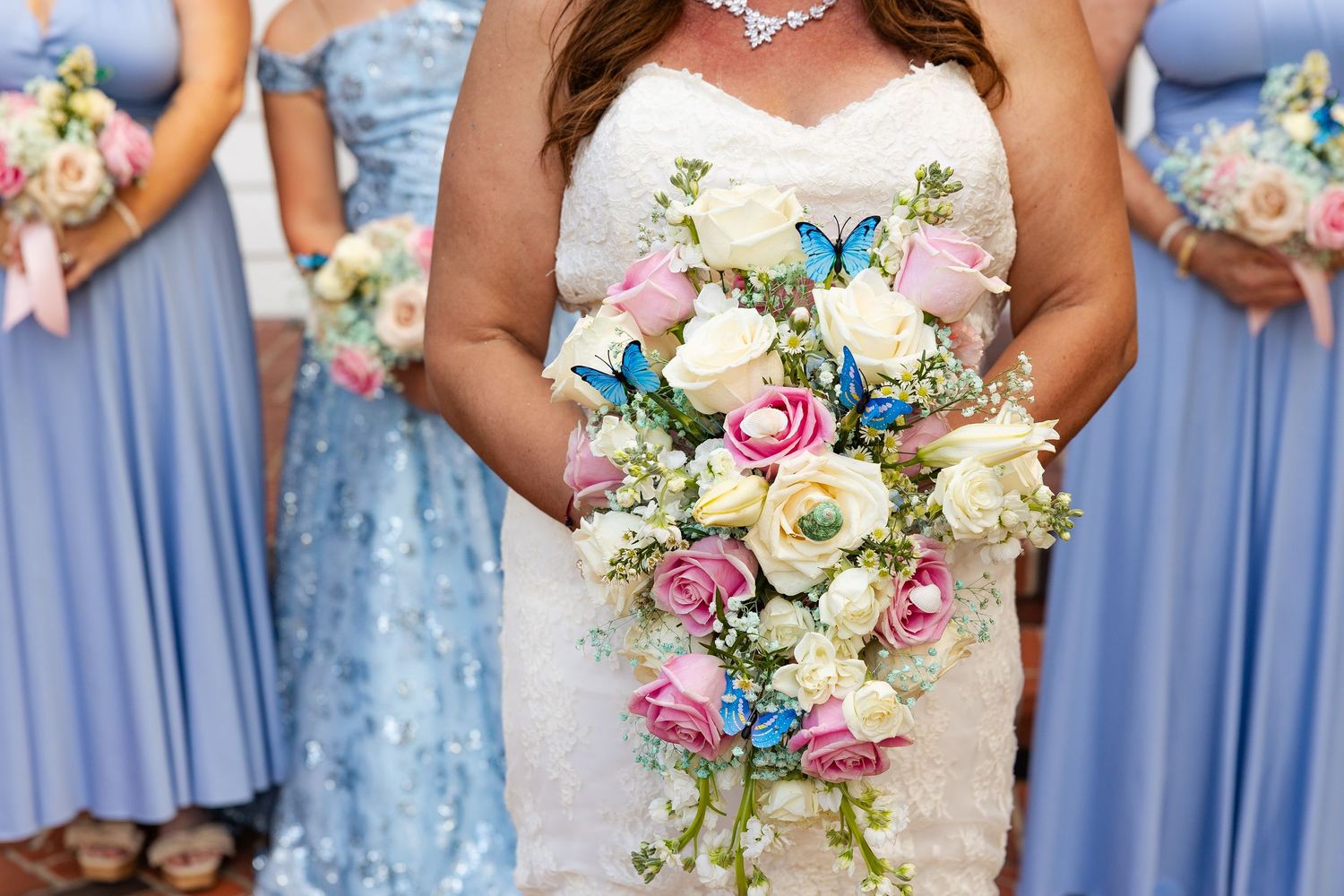 Bride holds stunning cascading bouquet of roses with blue butterflies, flanked by blue-gowned bridesmaids.