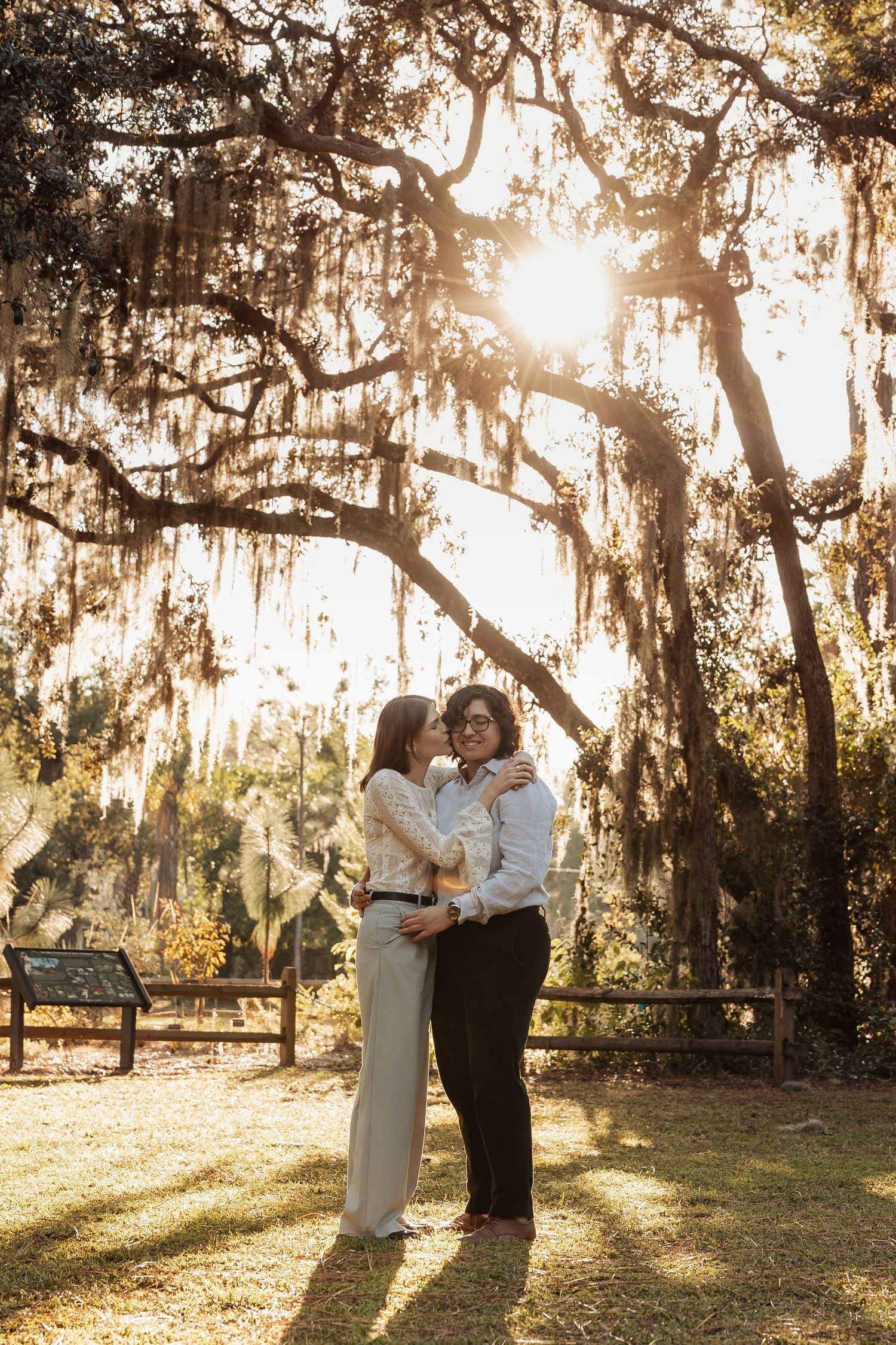 Two women sharing a kiss under large oak trees with Spanish moss in golden sunset light.