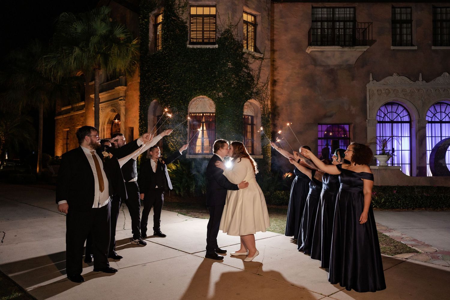 Bride and groom share a sparkler send-off with wedding party outside an illuminated historic venue at night.