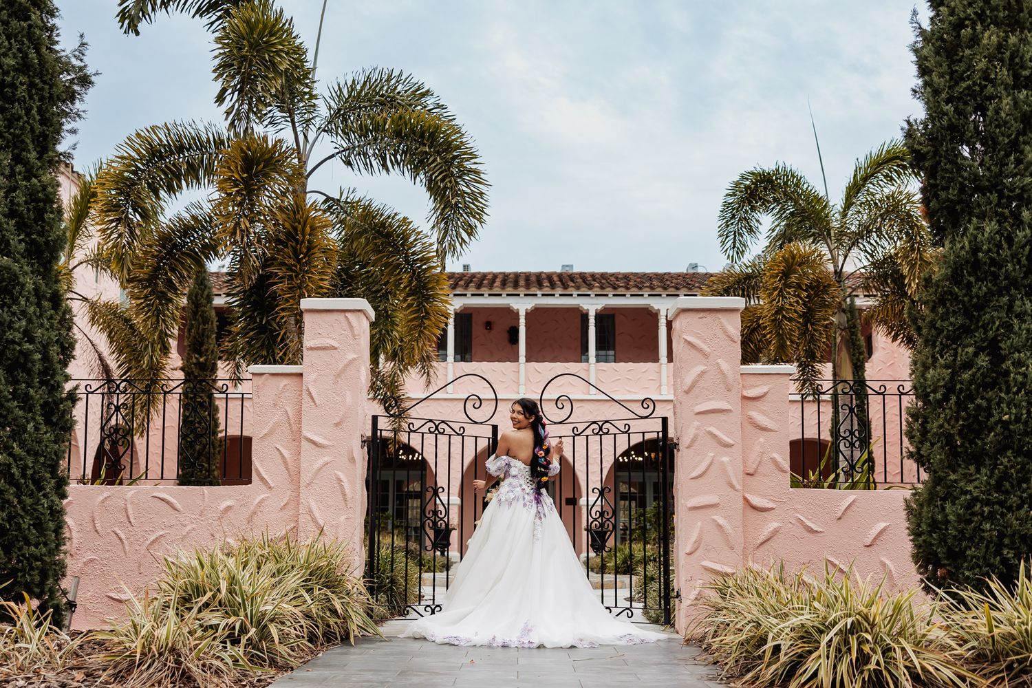 A figure in a white wedding gown stands before an elegant pink Mediterranean-style mansion with palm trees.