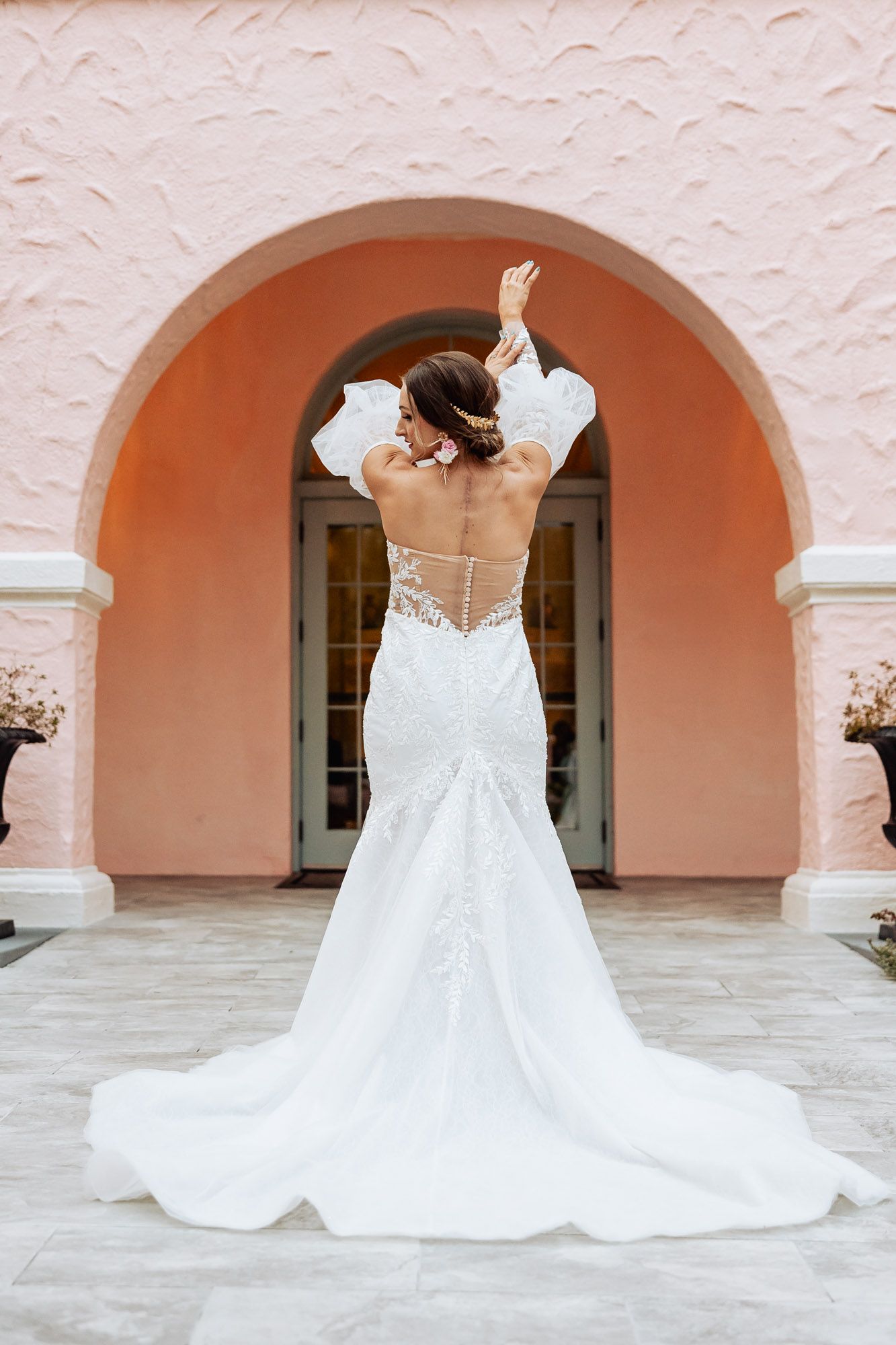 Bride in backless lace mermaid gown raises arms gracefully beneath pink arched entrance with train flowing.
