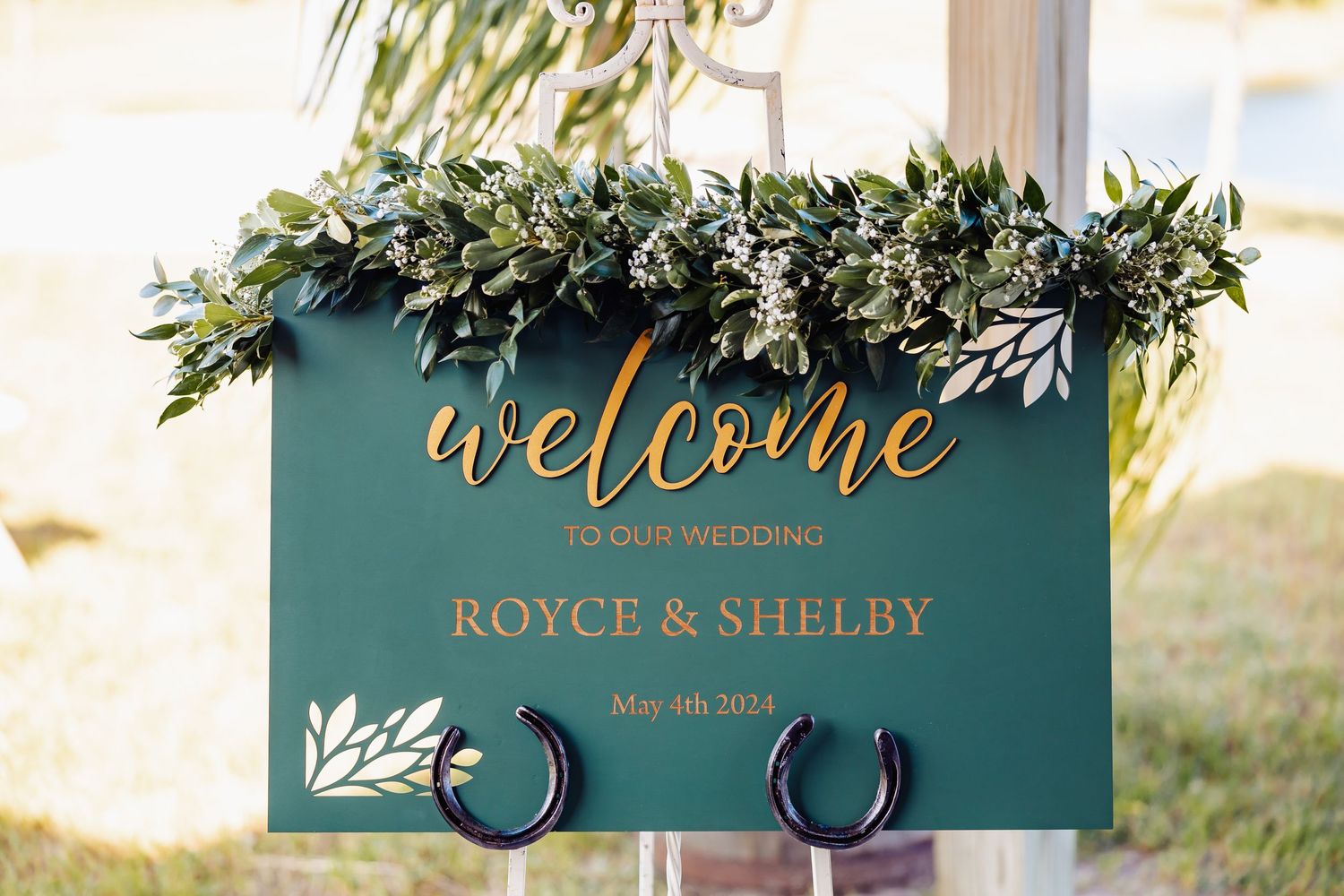 Elegant teal wedding welcome sign decorated with greenery and gold lettering.