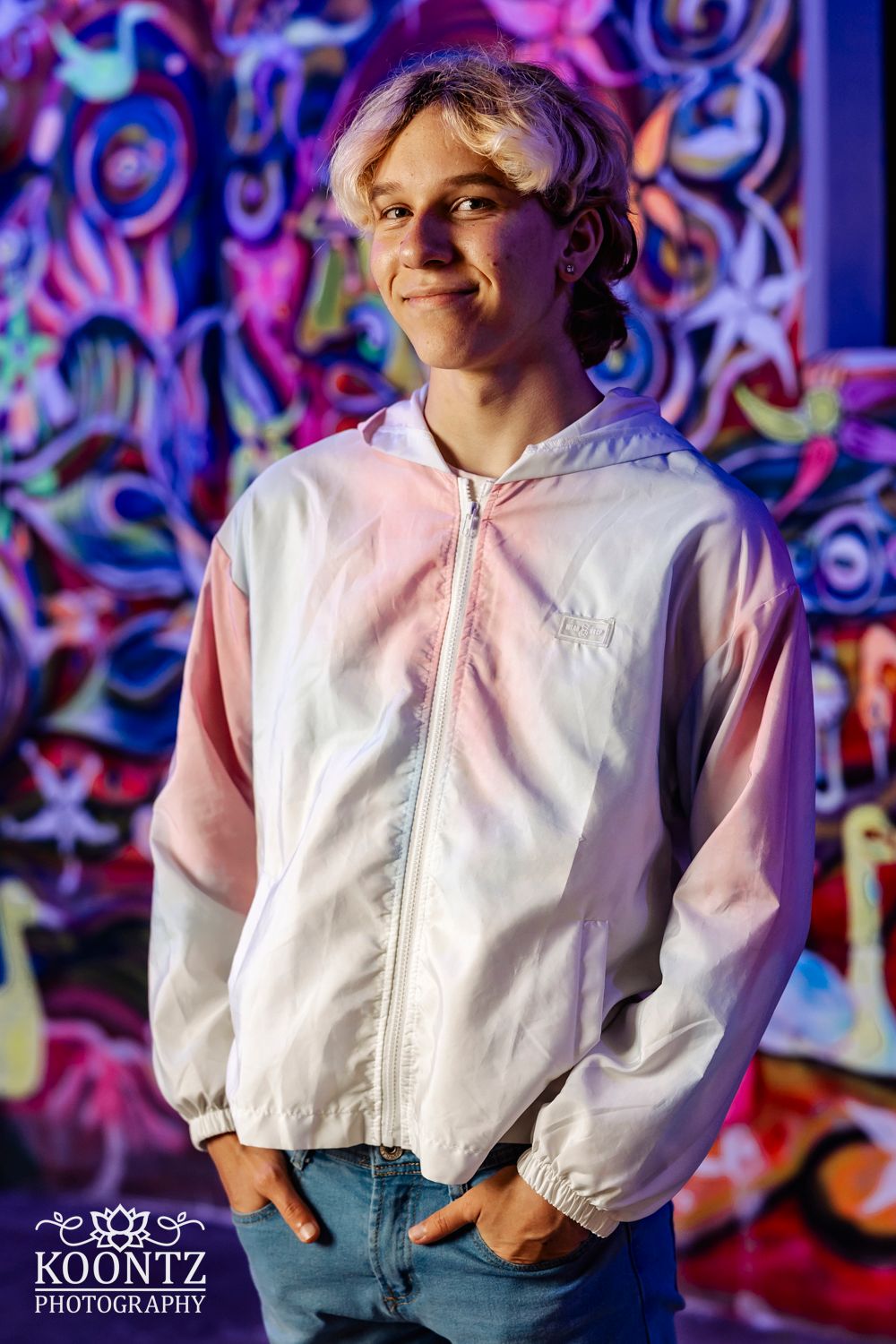 Portrait against vibrant neon graffiti wall wearing white and pink windbreaker jacket.