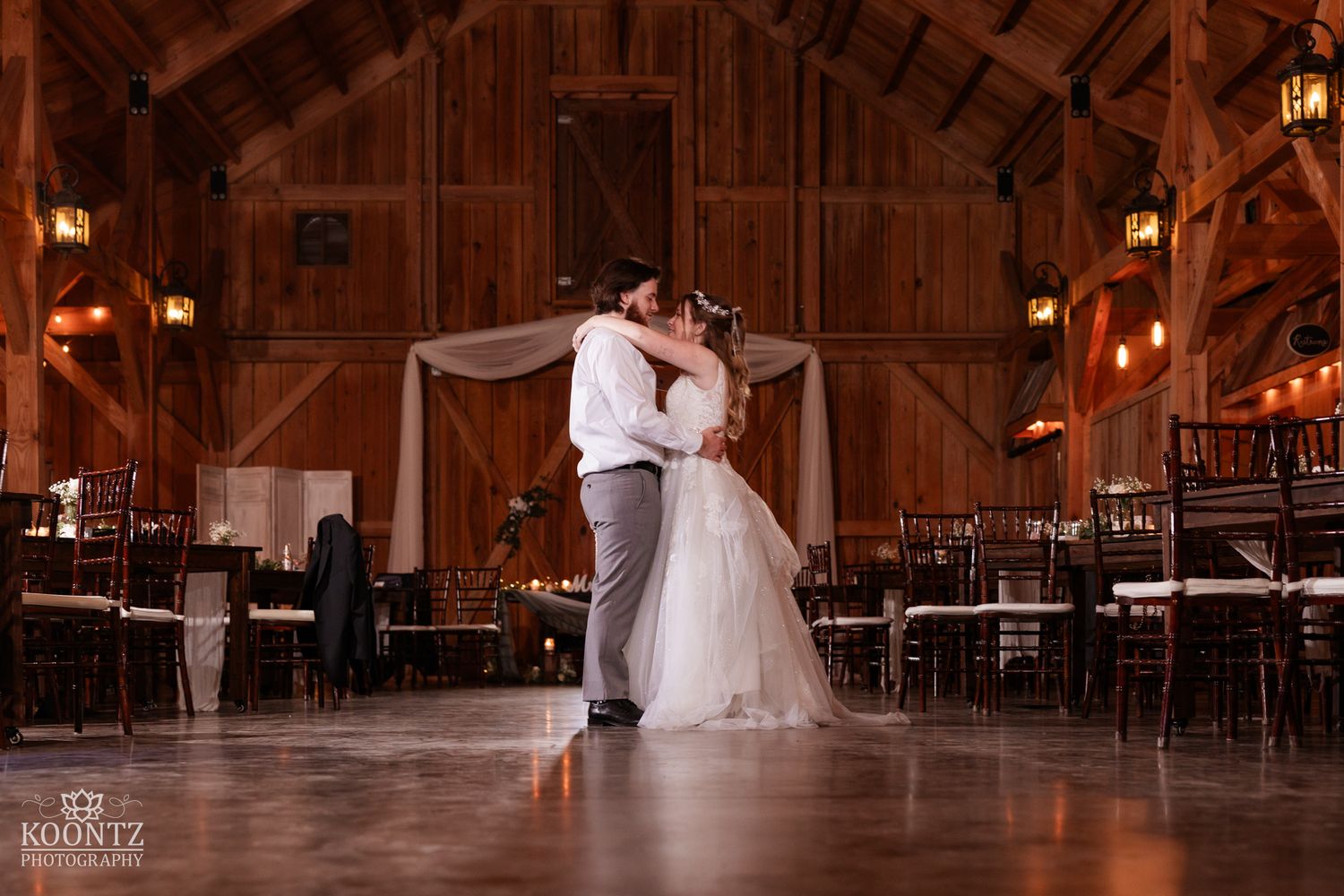 Bride and groom share first dance in elegant rustic barn wedding venue with warm ambient lighting.