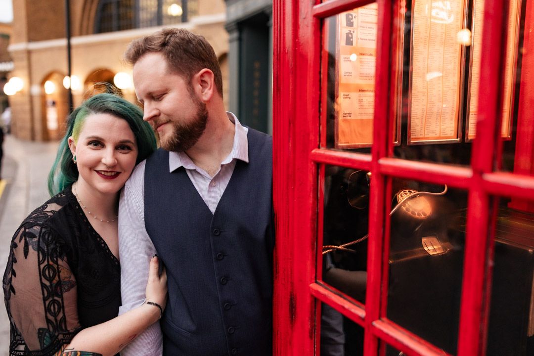 Couple posing together beside a classic red British telephone box on a city street.