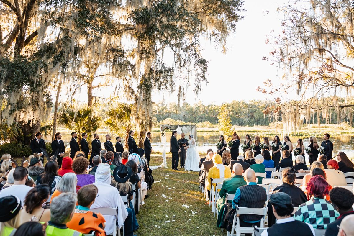 Outdoor wedding ceremony under Spanish moss-draped trees with guests seated by a lake.