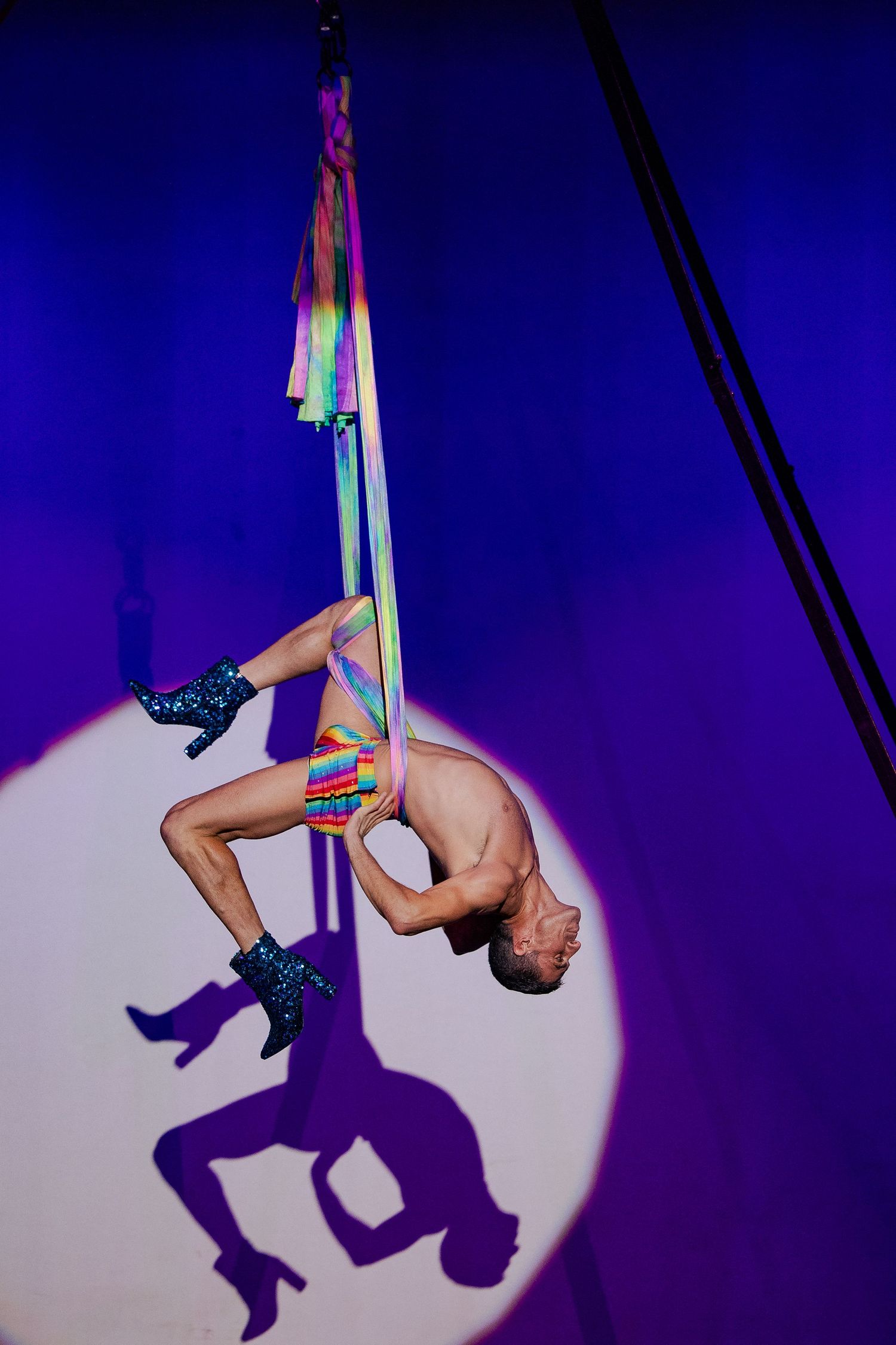Male aerialist in rainbow shorts performing inverted pose on aerial silks under purple spotlight.