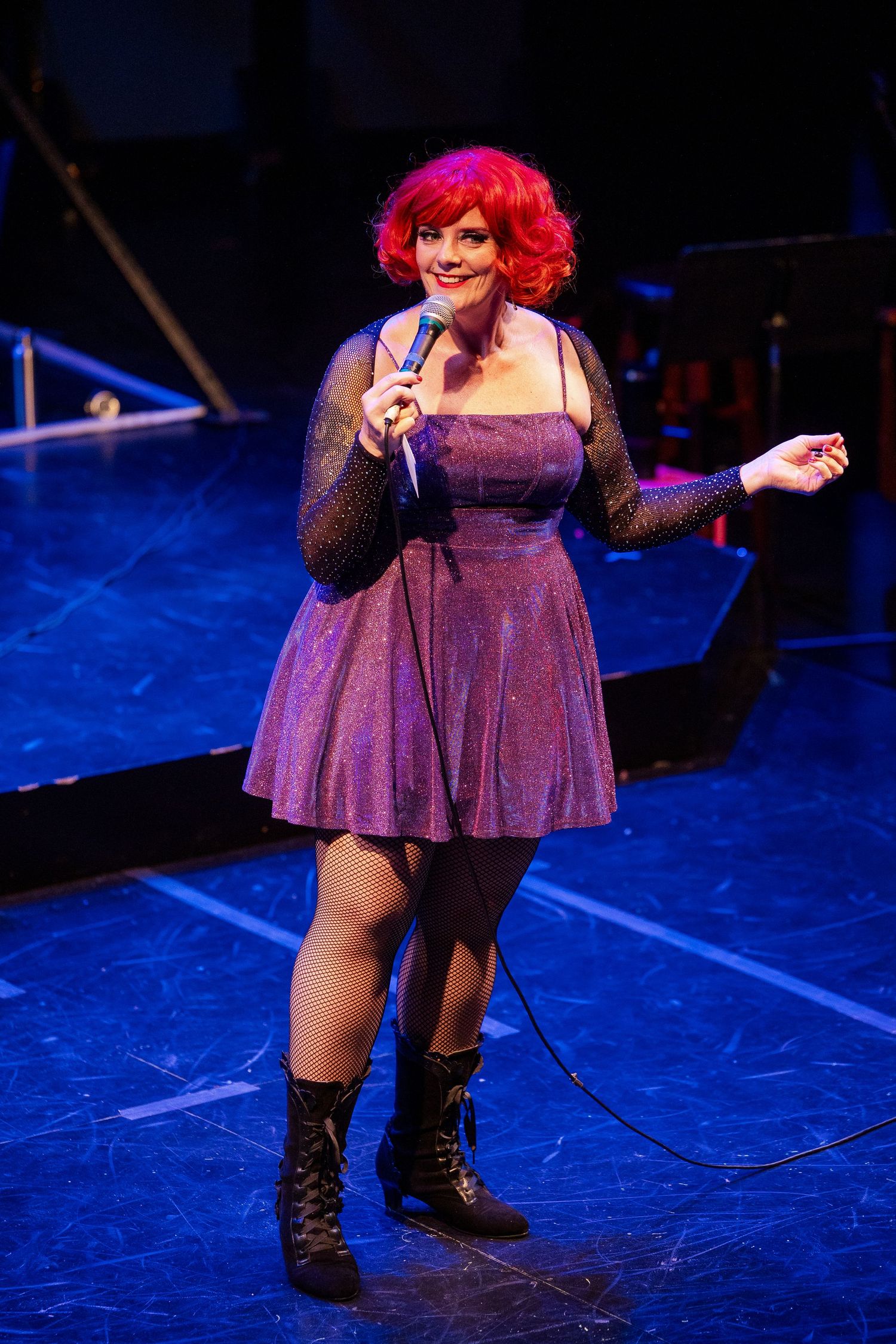 Red-haired female performer in purple sparkle dress singing into microphone on a blue-lit stage.
