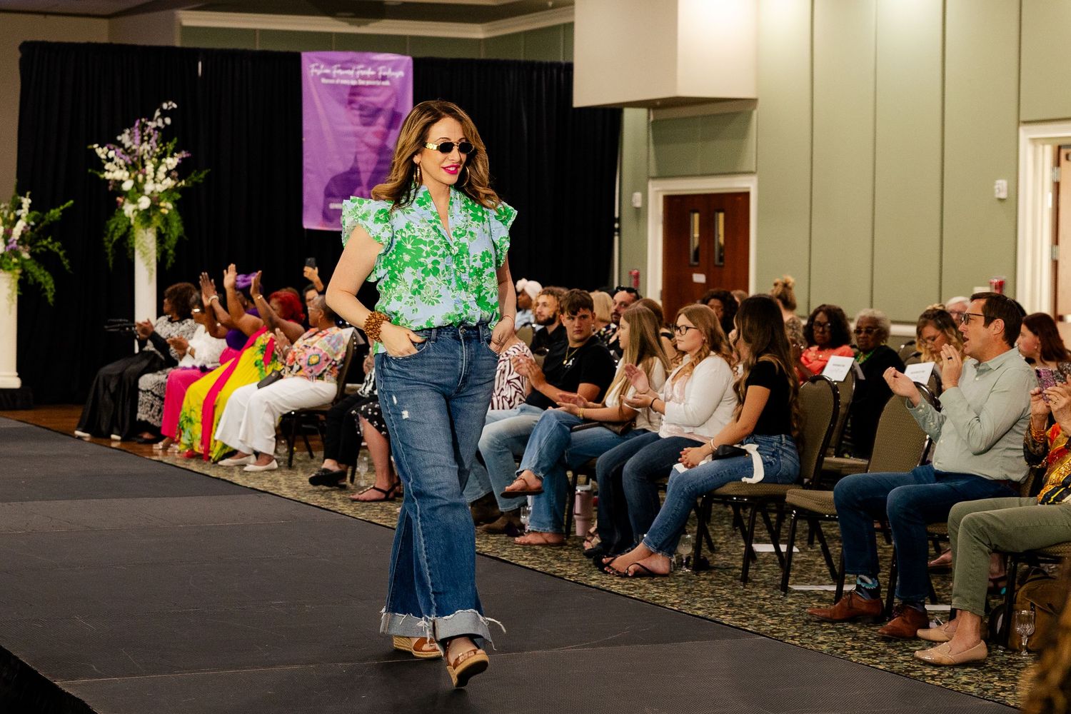 Model walks runway in green floral top and wide-leg jeans at fashion show.