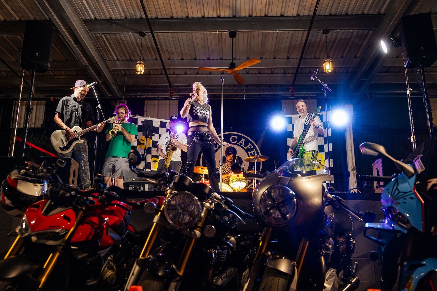A full band performs on a stage surrounded by motorcycles in what appears to be a garage venue at night.