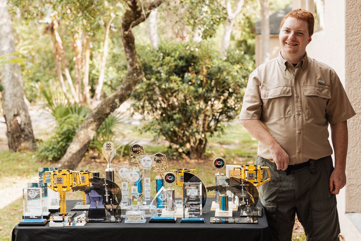 A smiling young man in khaki stands proudly beside a table displaying trophies and awards outdoors.