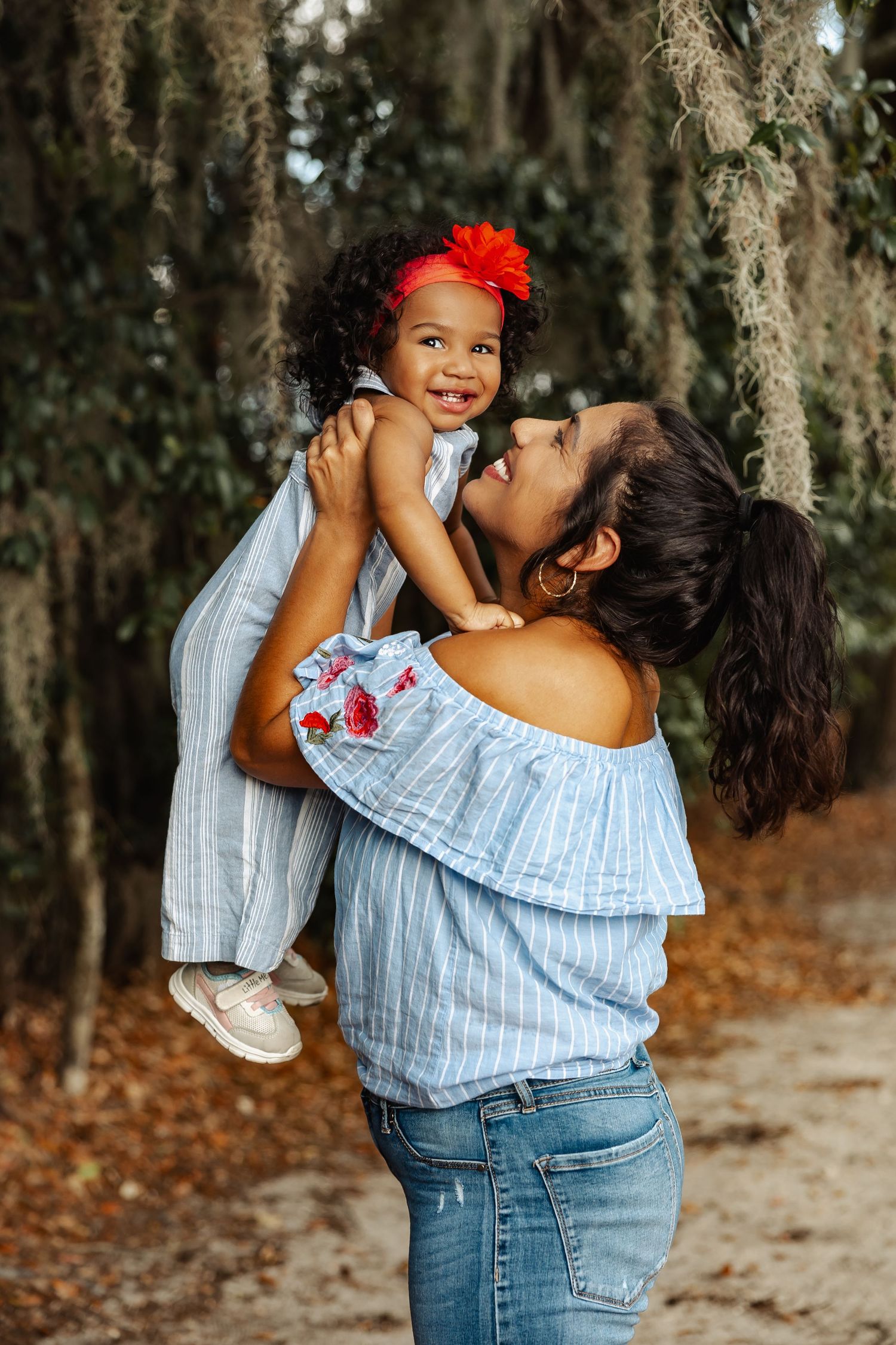 A mother lifts her laughing toddler daughter with a red bow in the air outdoors.