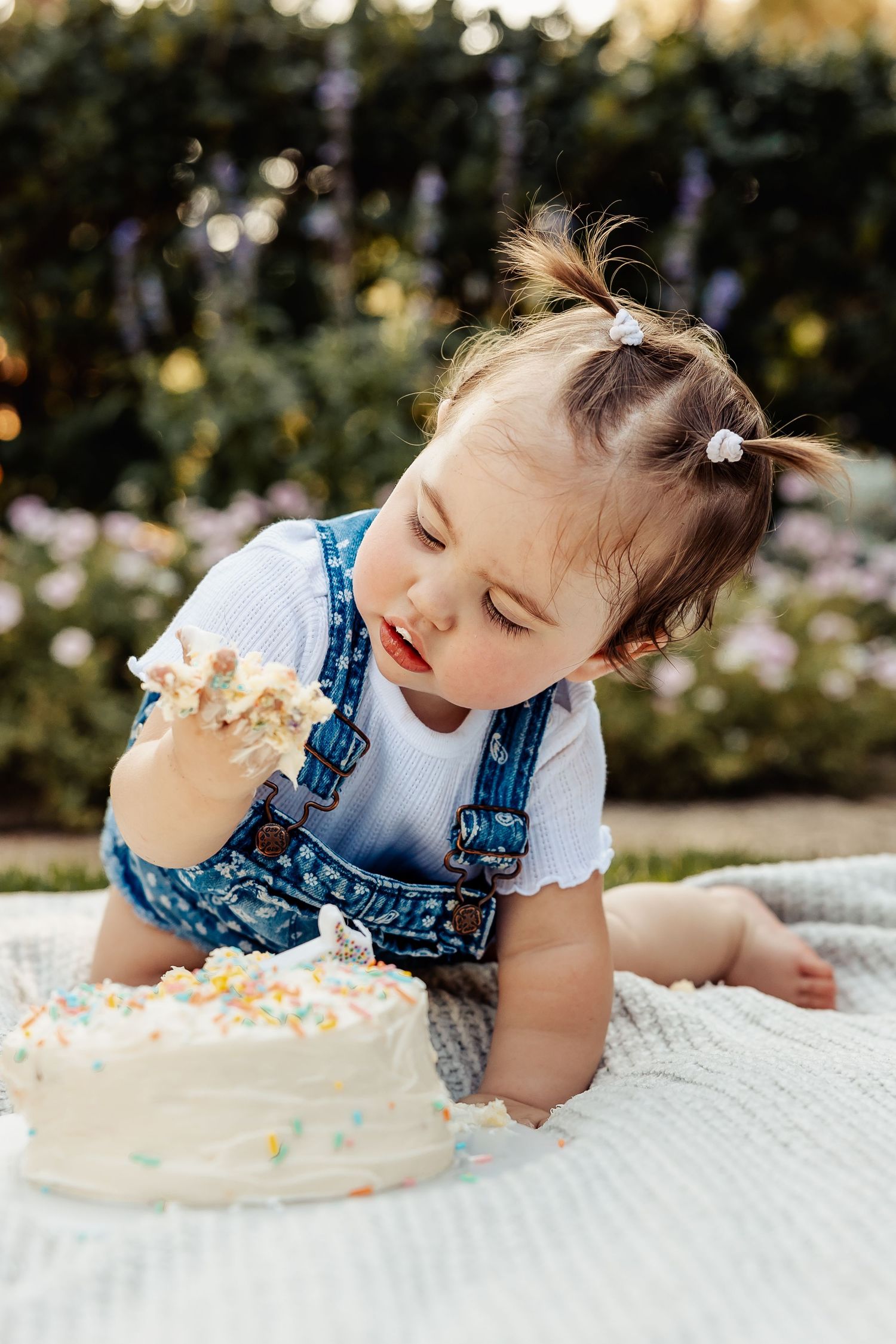 Baby girl in denim overalls grabbing handful of white frosting from birthday smash cake outdoors.