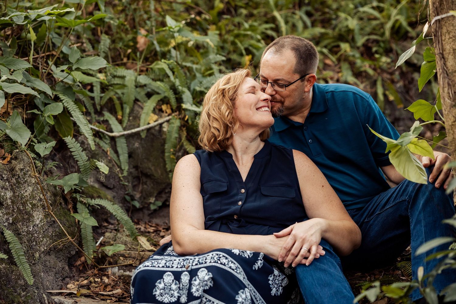 Romantic couple sitting together in a lush garden, leaning in for a kiss surrounded by green plants.