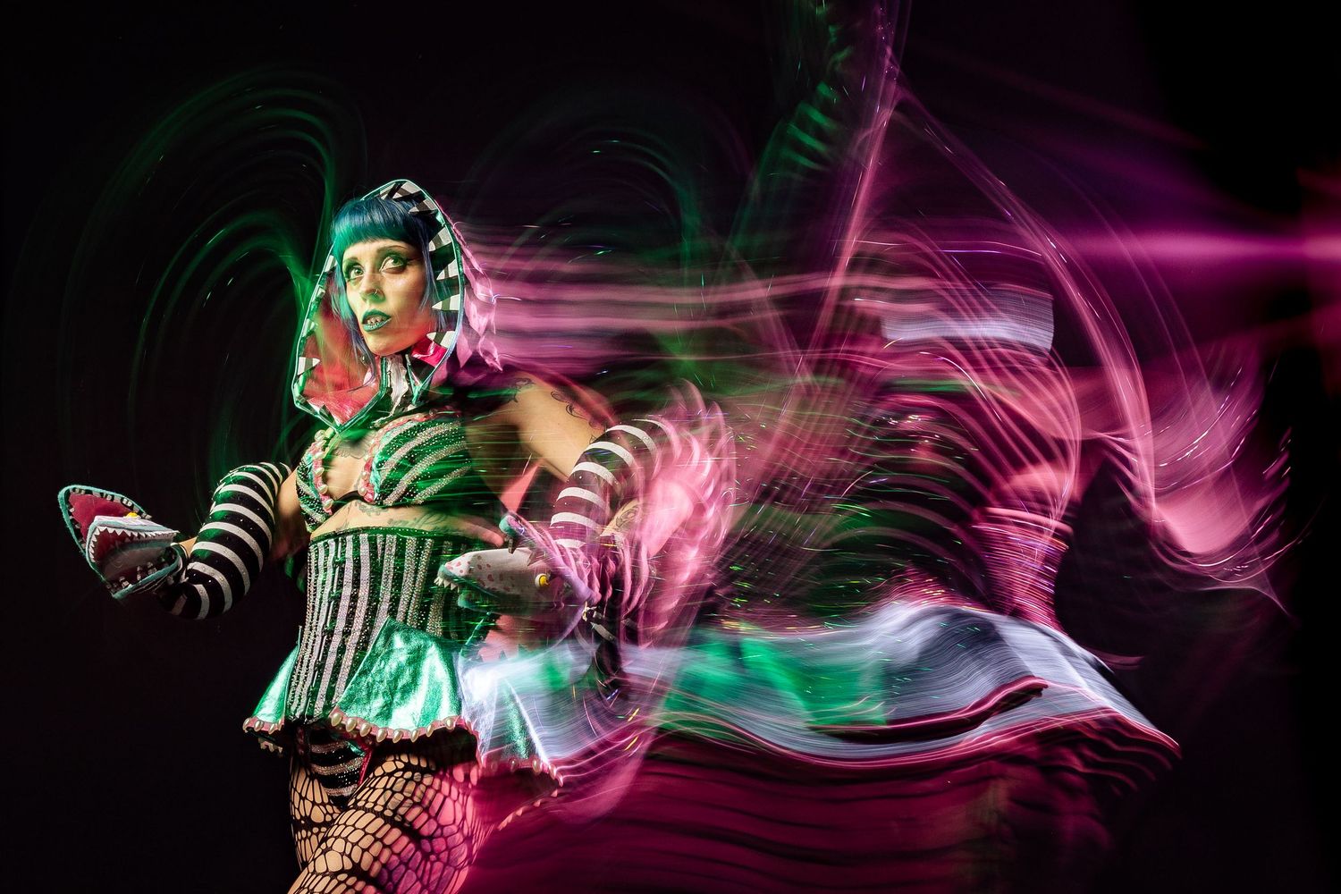 A vibrant performer in green costume with light painting trails in pink and green against a dark background.