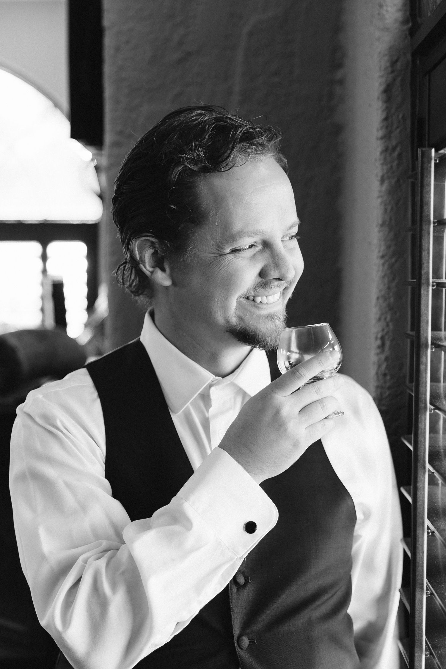 Handsome groom in a vest and dress shirt smiling while holding a glass of whiskey in black and white photo.
