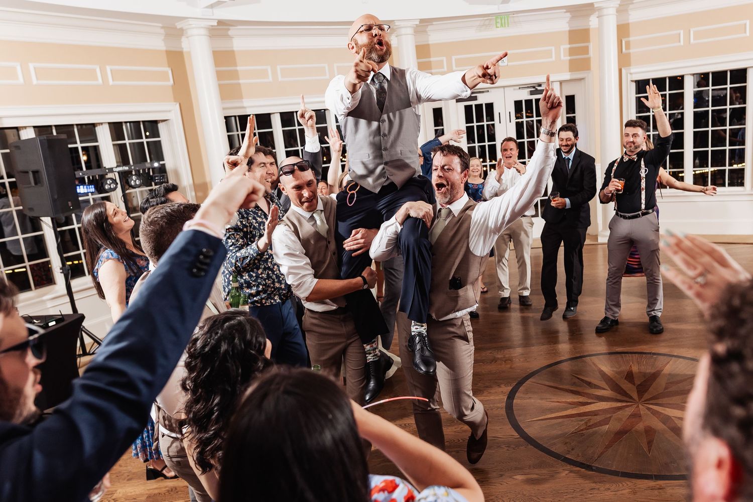 Groom lifted by groomsmen on dance floor while guests celebrate at lively wedding reception.