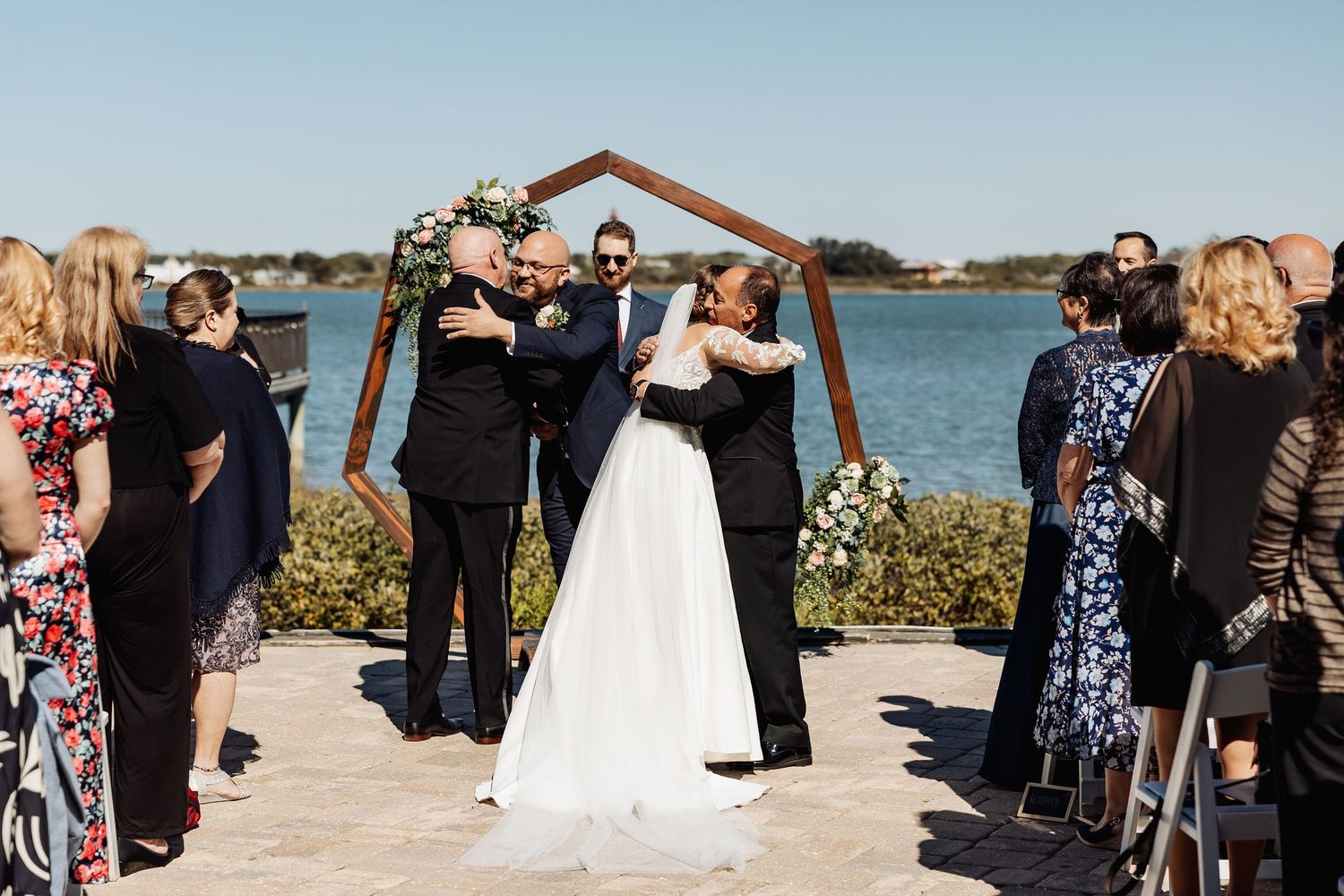 Bride and groom embrace family members at outdoor waterfront ceremony beneath a geometric wooden arch.