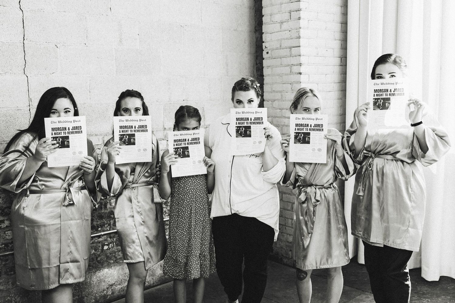 Bridesmaids in robes holding newspaper wedding announcements covering their faces in black and white photo.