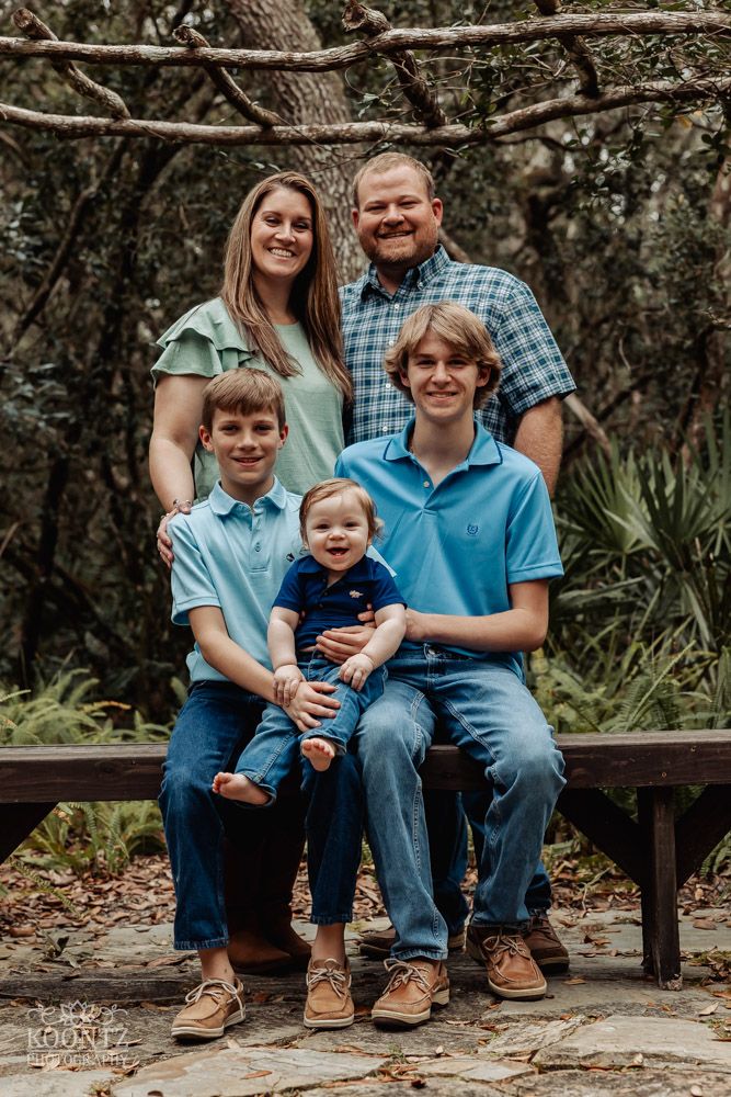 Family portrait of five people seated on a wooden bench in a natural outdoor setting.
