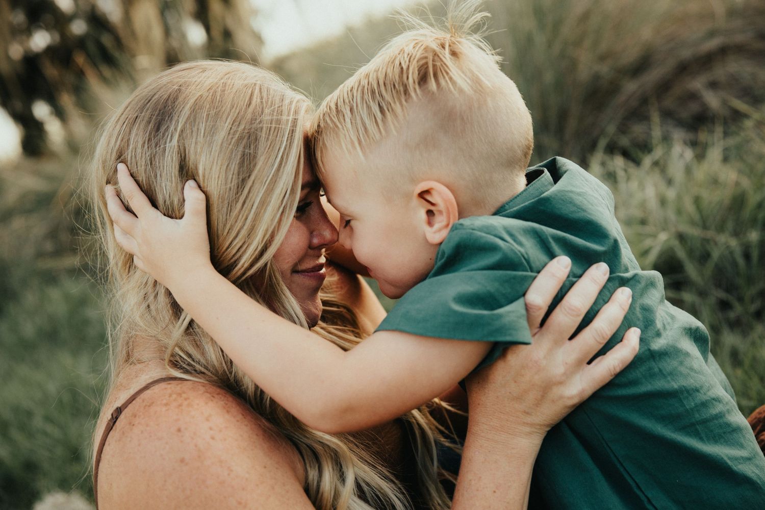 A tender moment of affection photographed outdoors with natural backlighting and mossy trees.