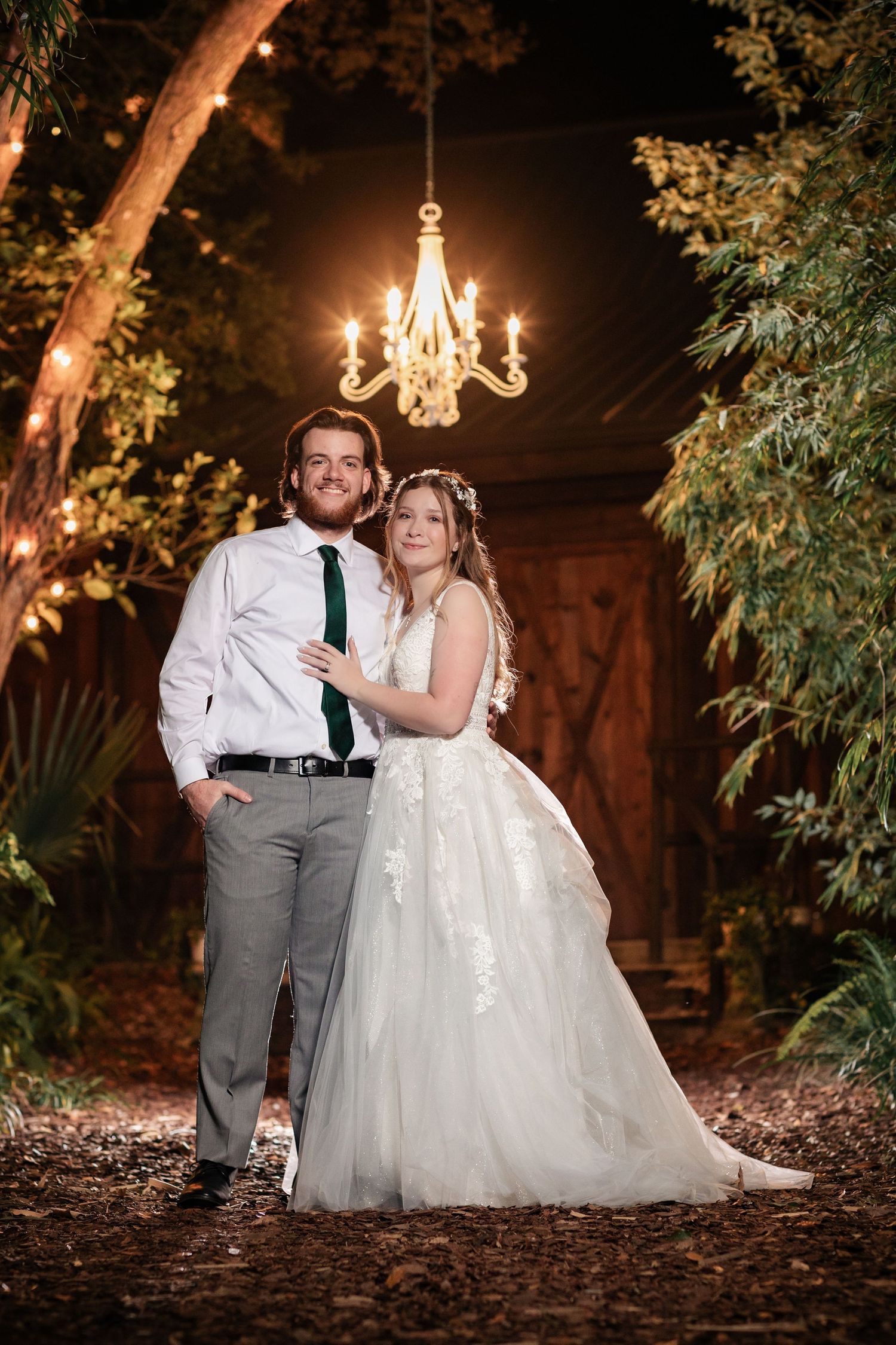 Evening portrait under hanging chandelier with rustic barn and forest backdrop.