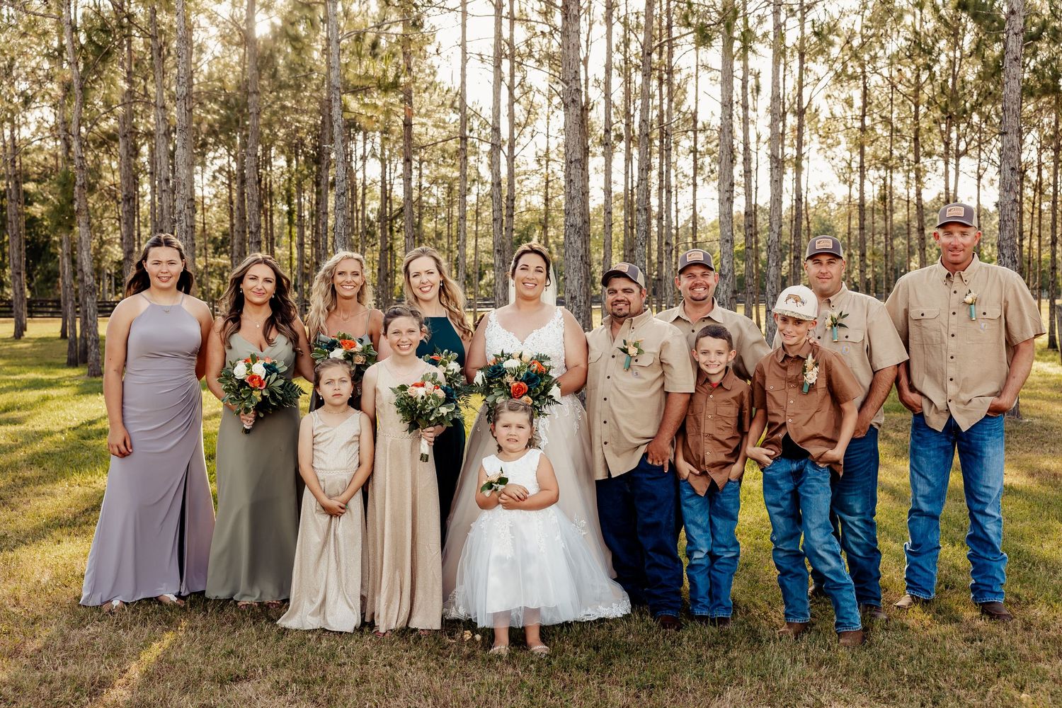 Wedding party poses together in a sunlit pine forest wearing mixed formal and casual attire.