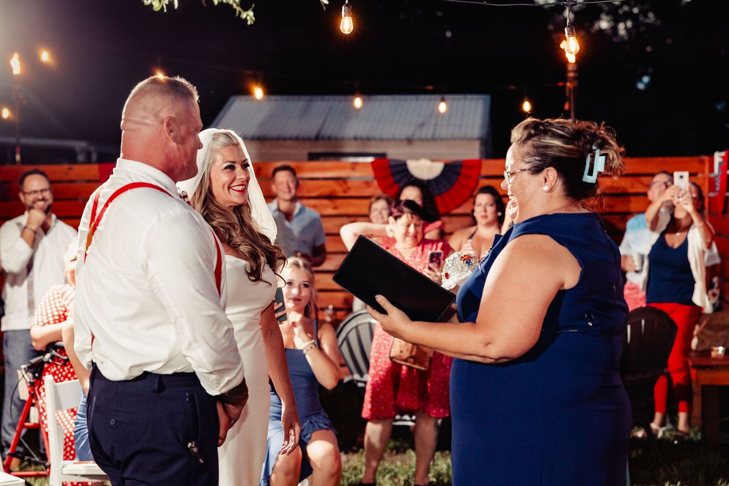 Couple exchanging vows at outdoor evening wedding ceremony while officiant reads from book with guests watching.