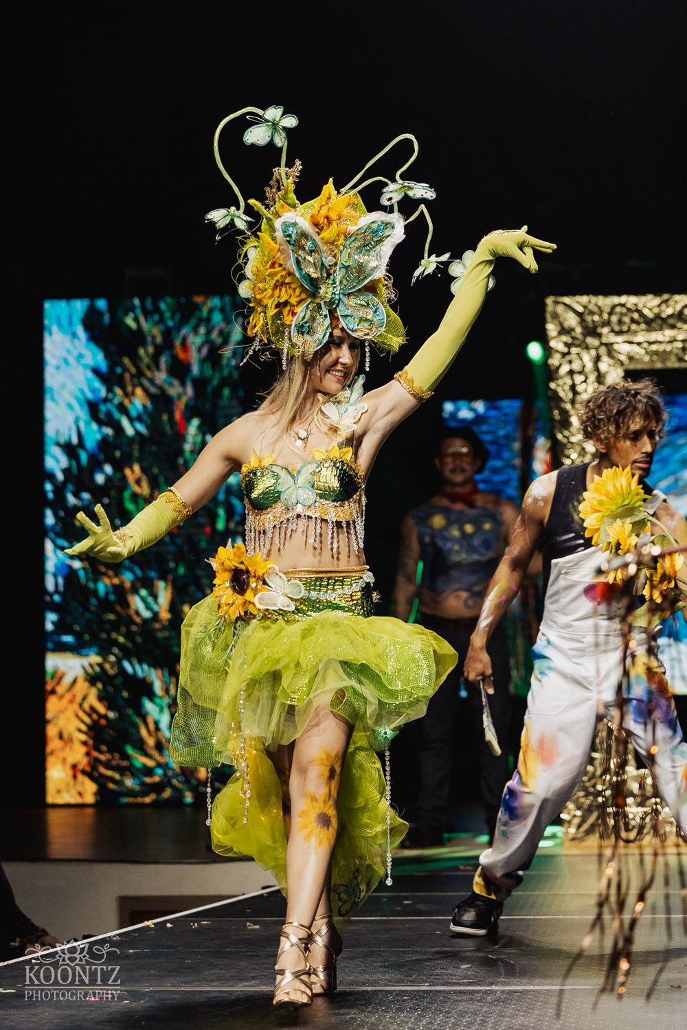 Dancer wearing yellow and green carnival costume with floral headpiece performing energetic dance routine on stage.