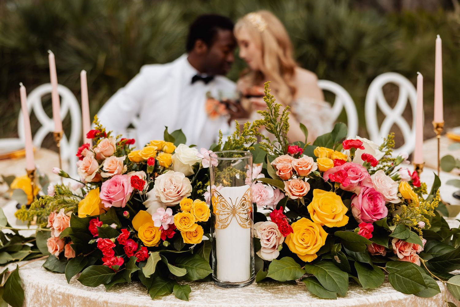 Colorful floral centerpiece with pink, yellow and coral roses adorns an elegant wedding table with white taper candles.
