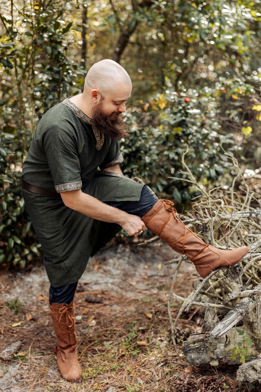 Person in dark clothing adjusts brown leather boots while standing in wooded area.