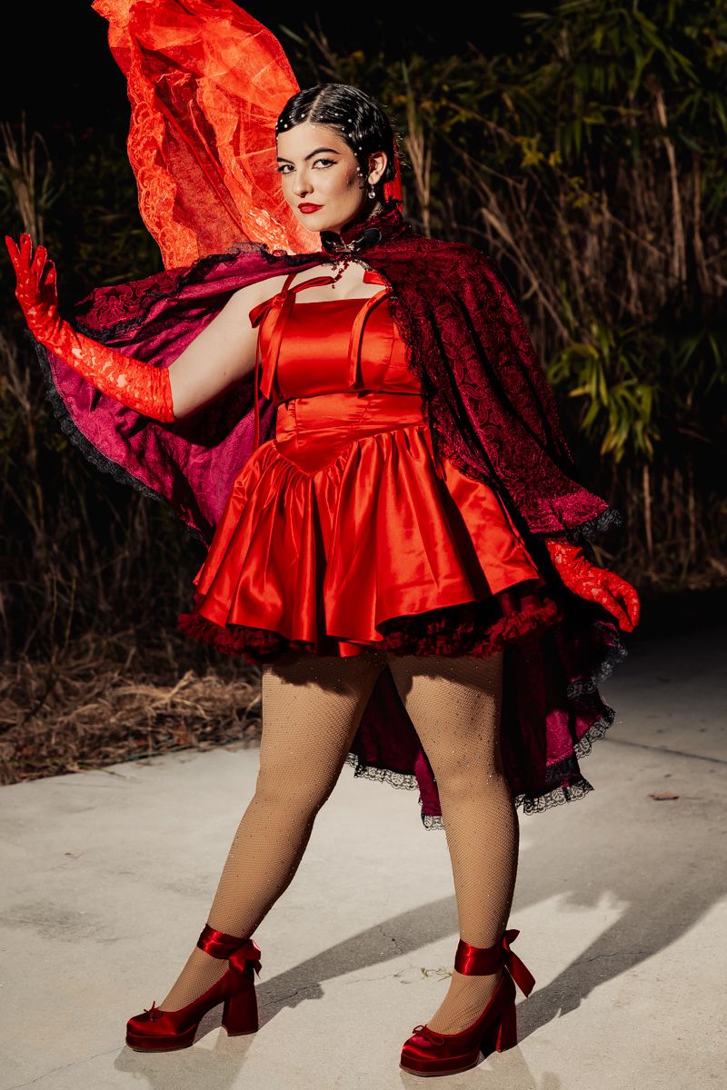 Woman in dramatic red satin mini dress with flowing lace cape and red heels posing outdoors.