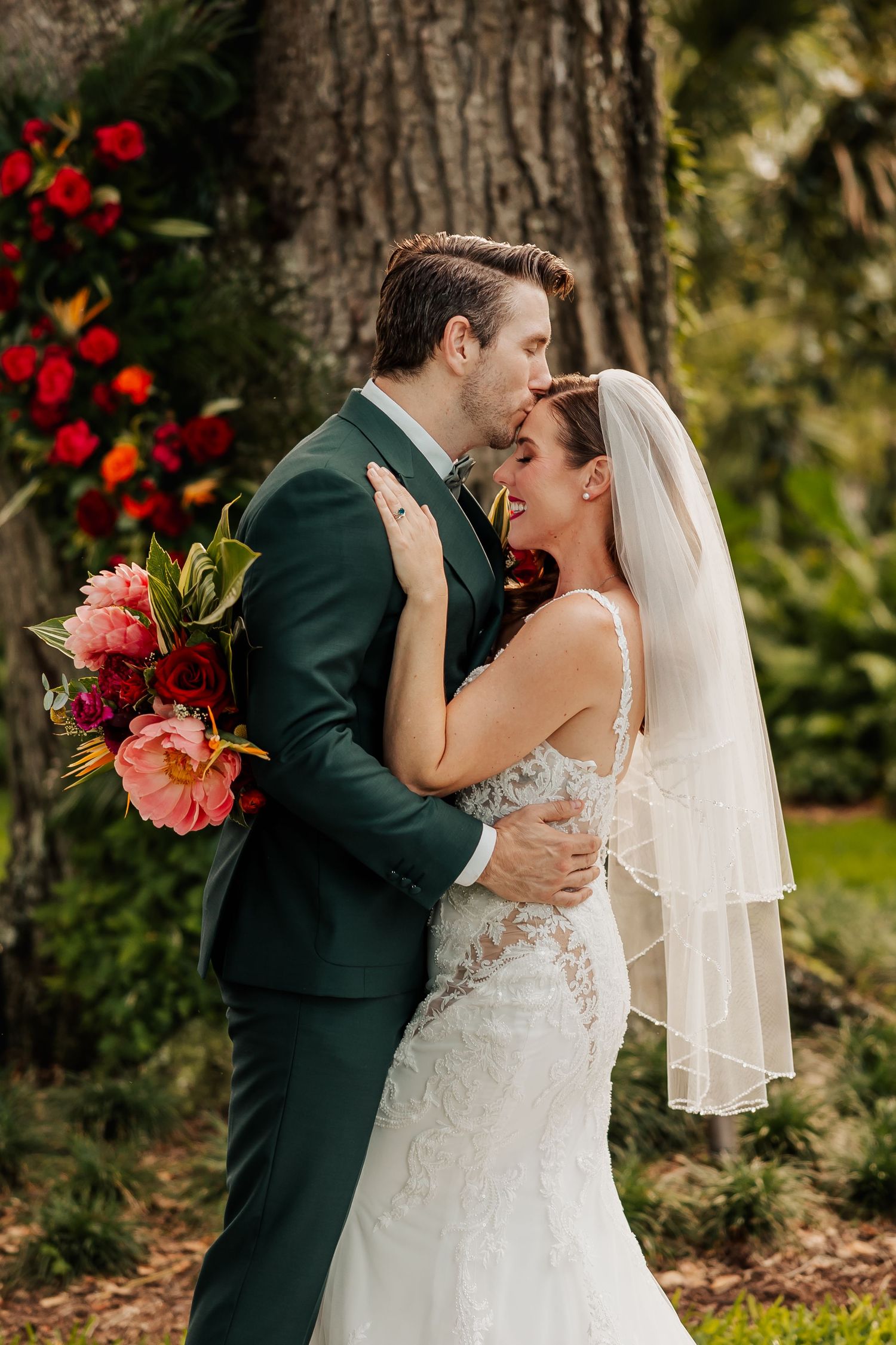 Laughing bride and groom embrace outdoors near large tree with vibrant red and pink floral decor.
