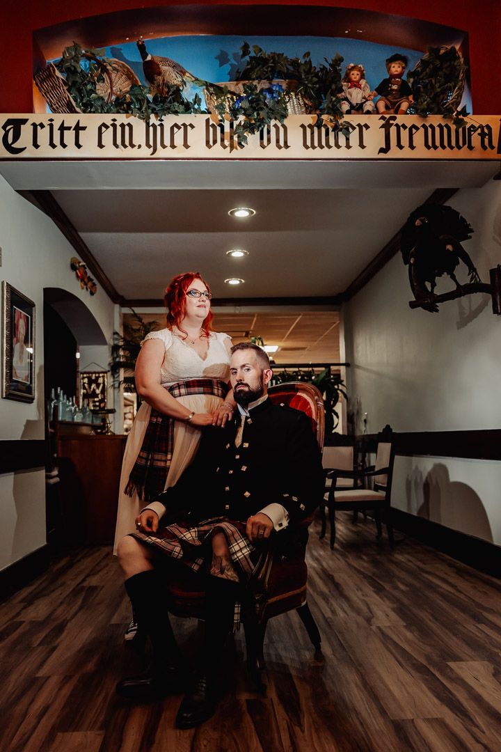 Couple posing dramatically indoors under German gothic script banner with kilt attire.