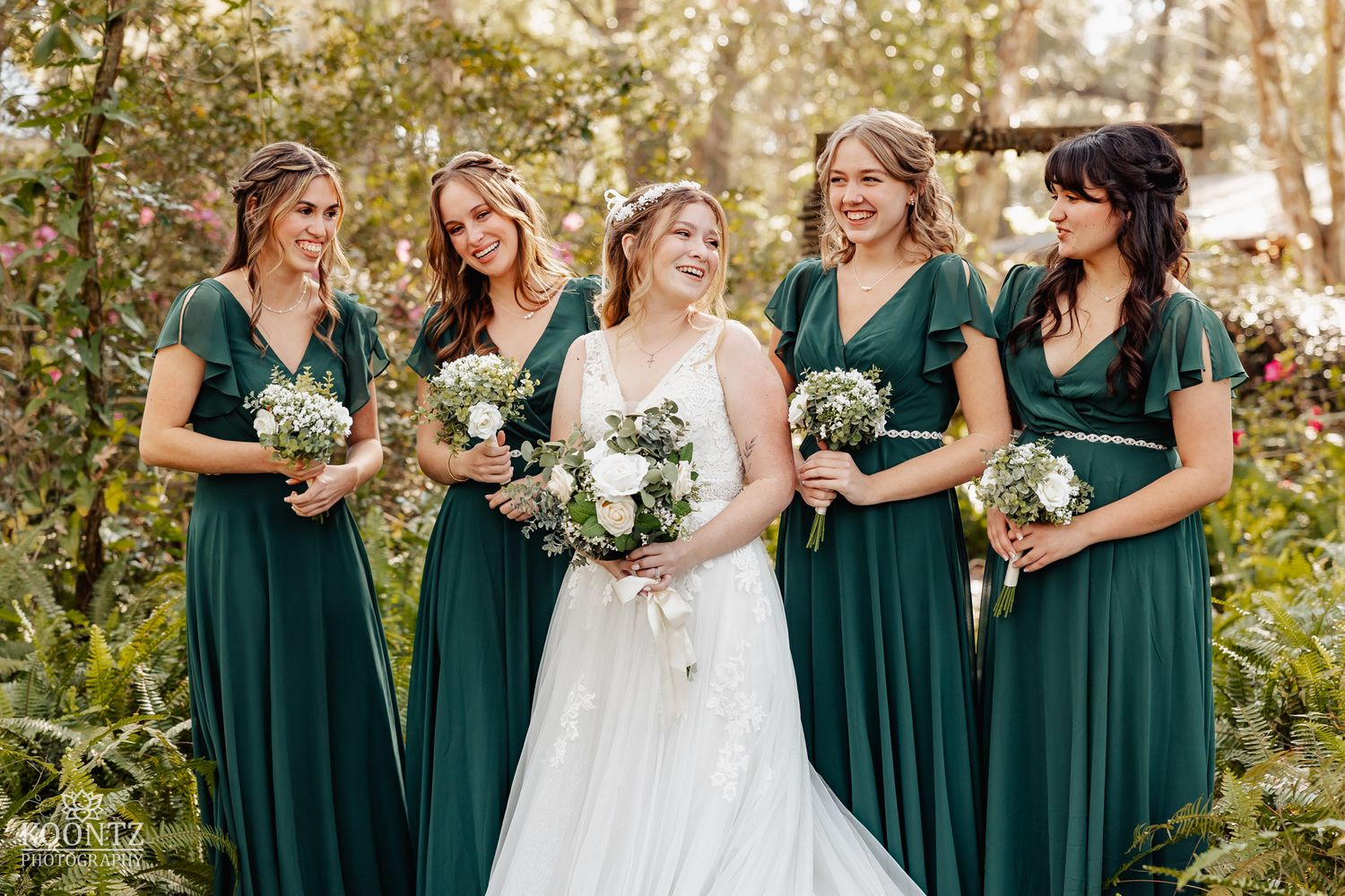 Bride and four bridesmaids in emerald green dresses smile together in a lush garden setting.