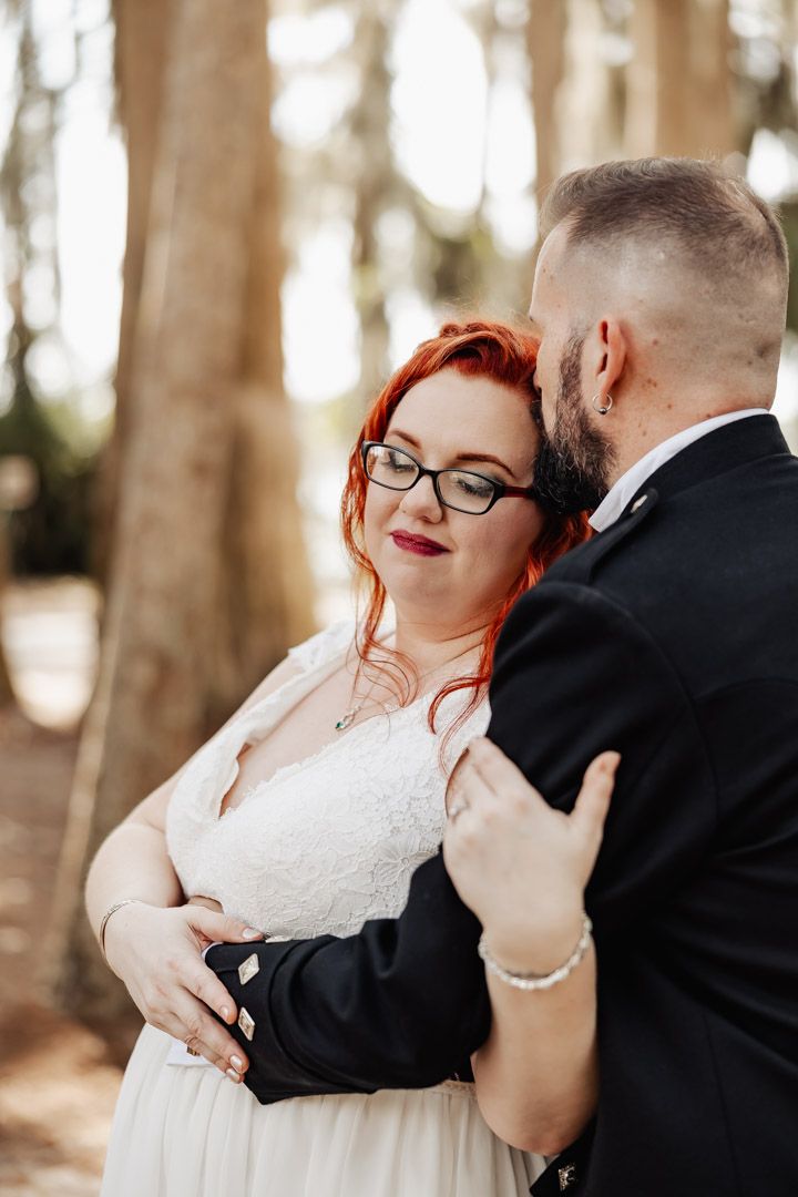 Bride with vibrant red hair and glasses embraced by groom in black jacket among tall trees.
