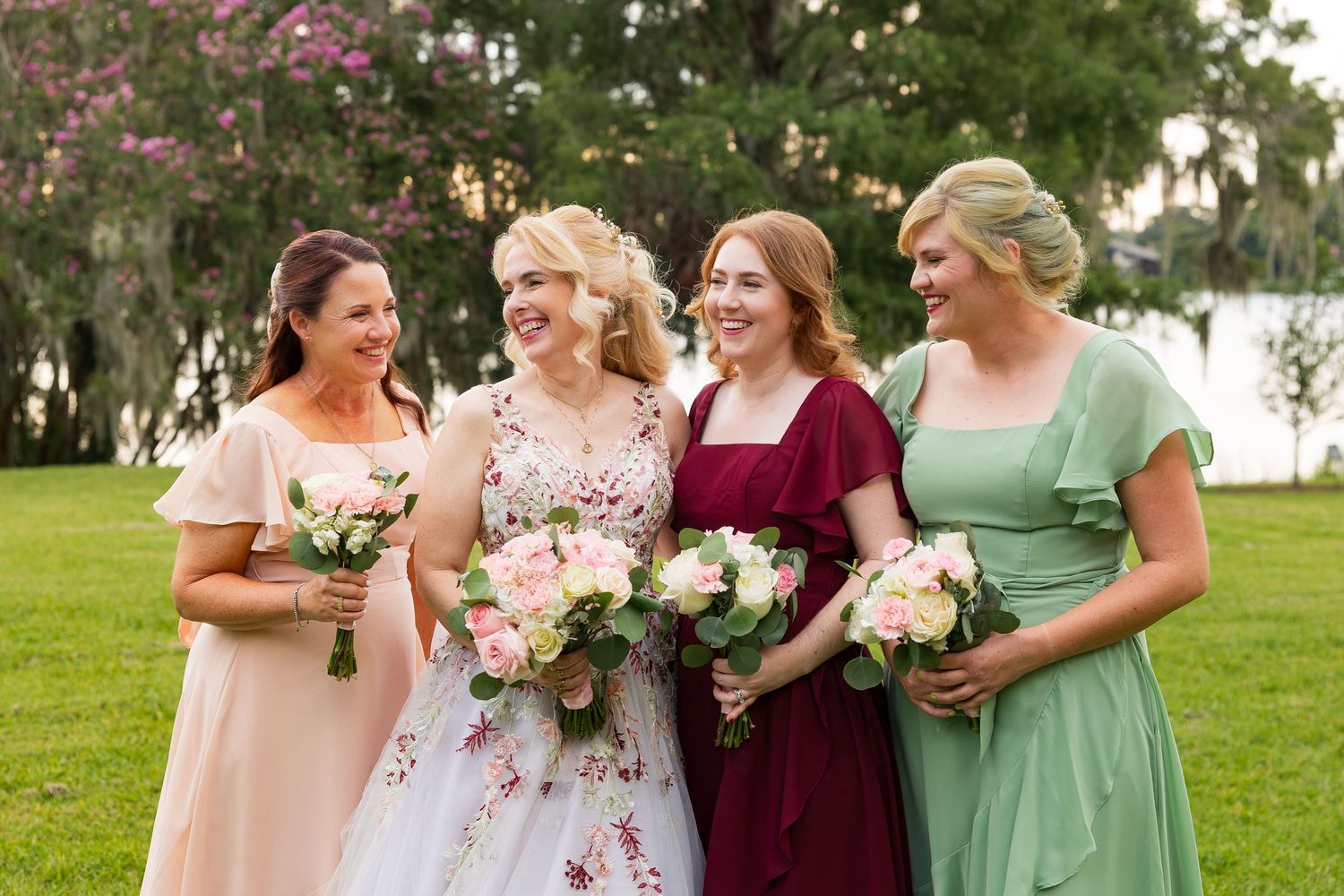 Bride and three bridesmaids in colorful dresses laugh together outdoors holding floral bouquets.
