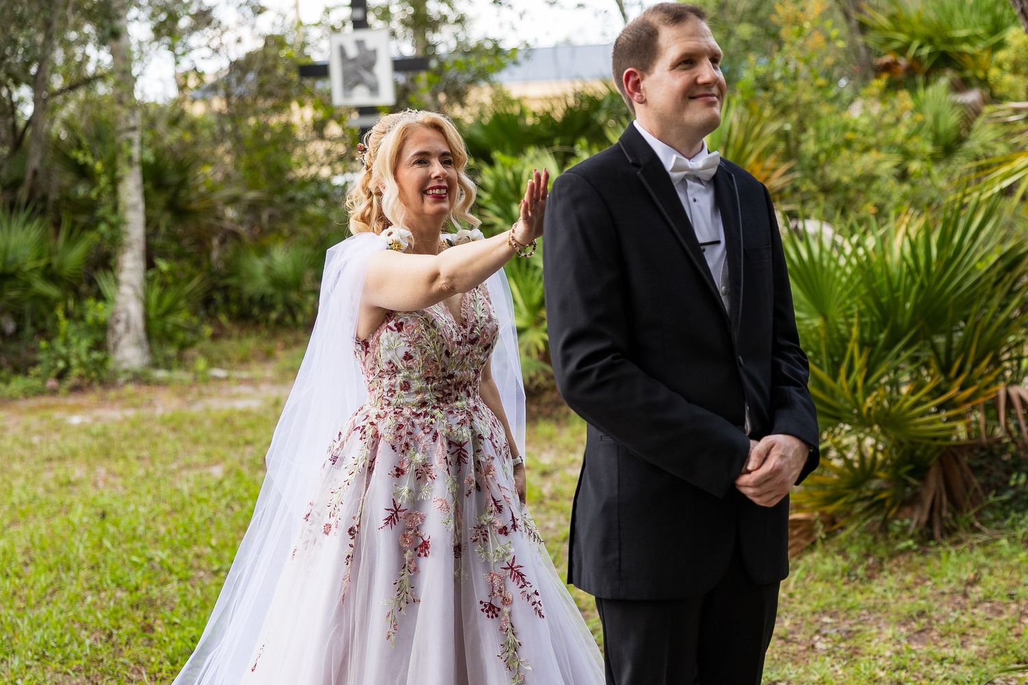 Emotional bride in floral wedding gown touching groom's face during outdoor first look reveal.