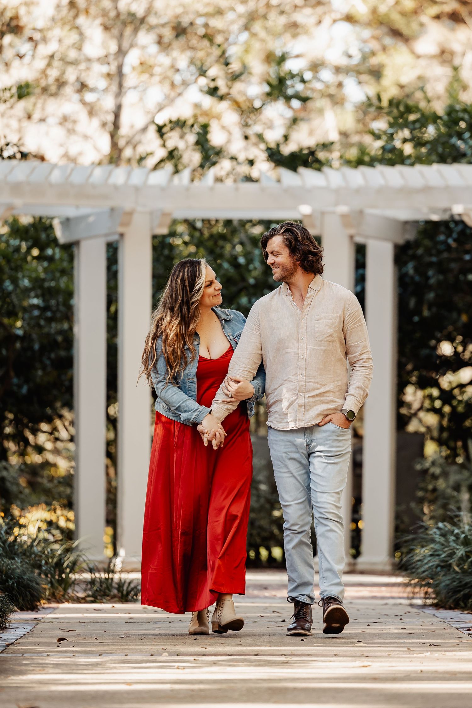 Couple walking hand-in-hand past white columns on a wooden path in a sunlit garden.