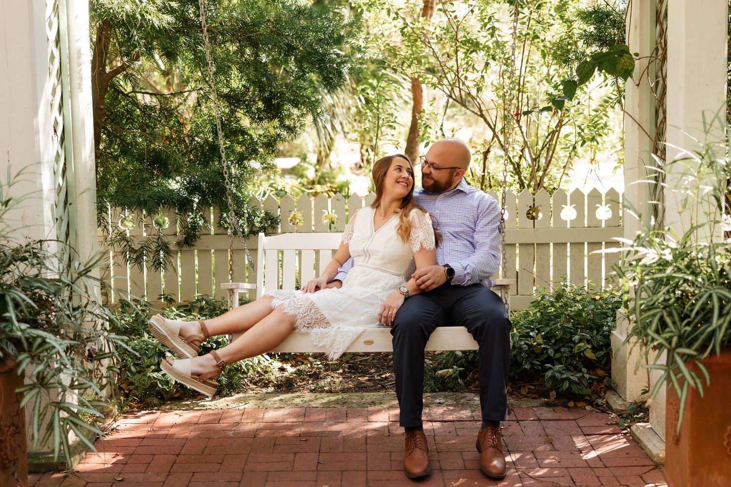Happy couple sitting together on white garden bench surrounded by green trees and plants.