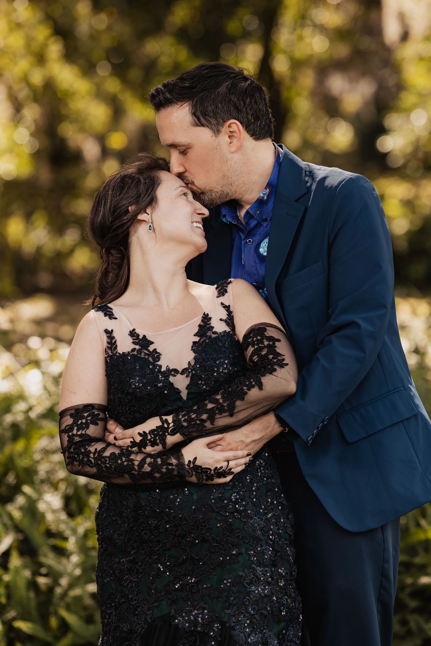 Groom in navy suit kisses laughing bride wearing black lace gown in sunlit garden wedding portrait.