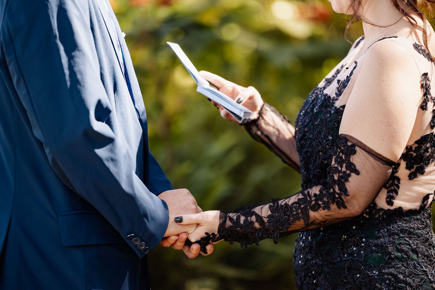 Bride in black lace gown holding vow booklet while exchanging vows with groom outdoors.