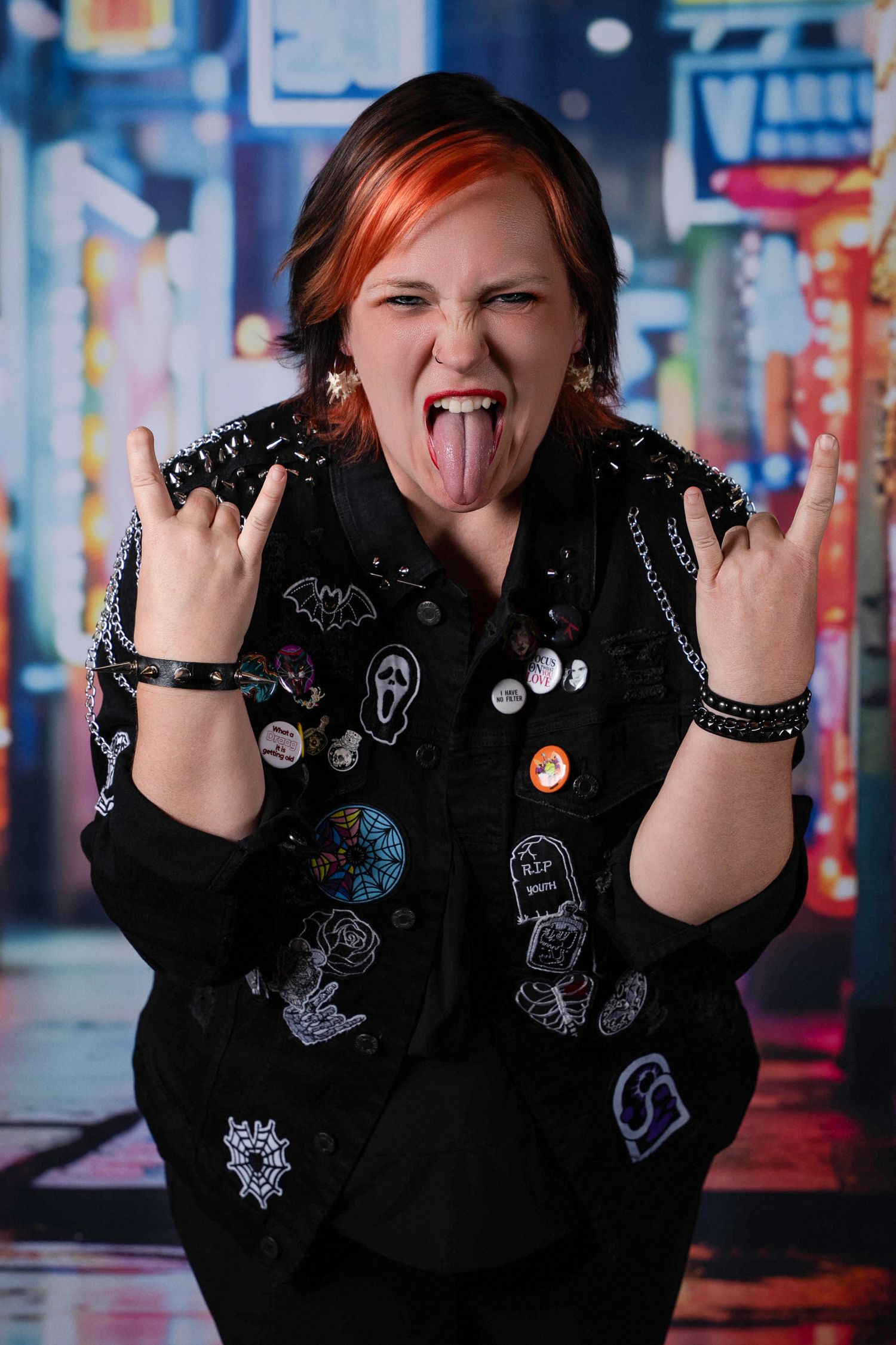 Punk woman with red hair making rock horns gesture, tongue out, wearing black jacket with pins and patches.