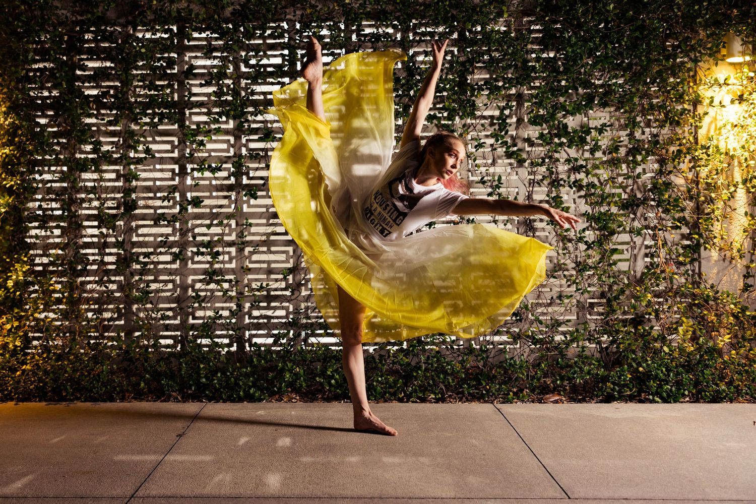 A barefoot dancer twirls a vibrant yellow fabric skirt against a lush ivy-covered wall outdoors at night.