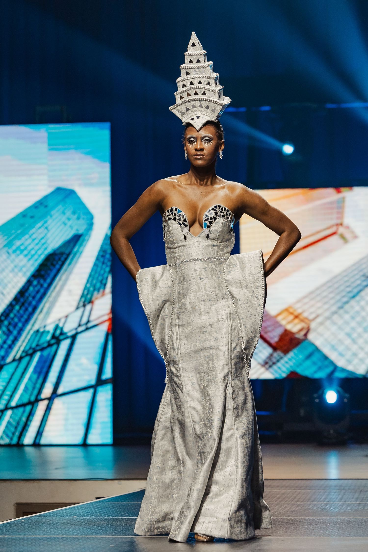 Model in silver strapless gown with tall tiered architectural white crown walks fashion runway.