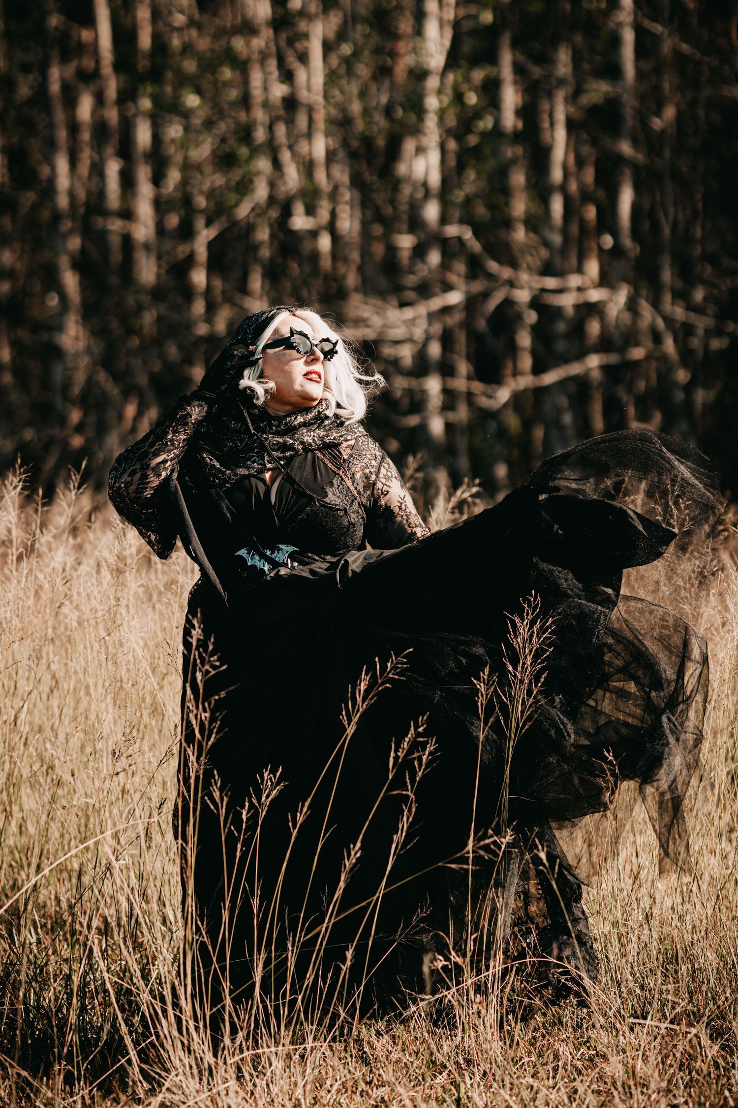 Woman in flowing black lace gown stands in autumn field with wind-swept fabric near pine forest.