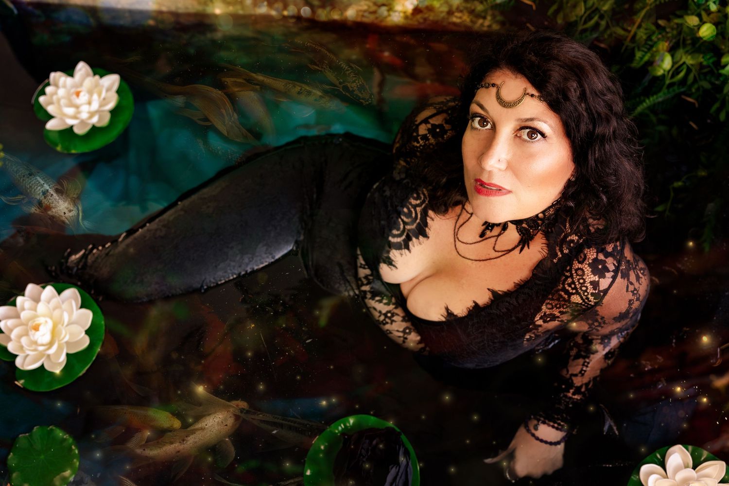 Dark-haired woman in black lace top sits beside lily pad pond in mystical garden scene.